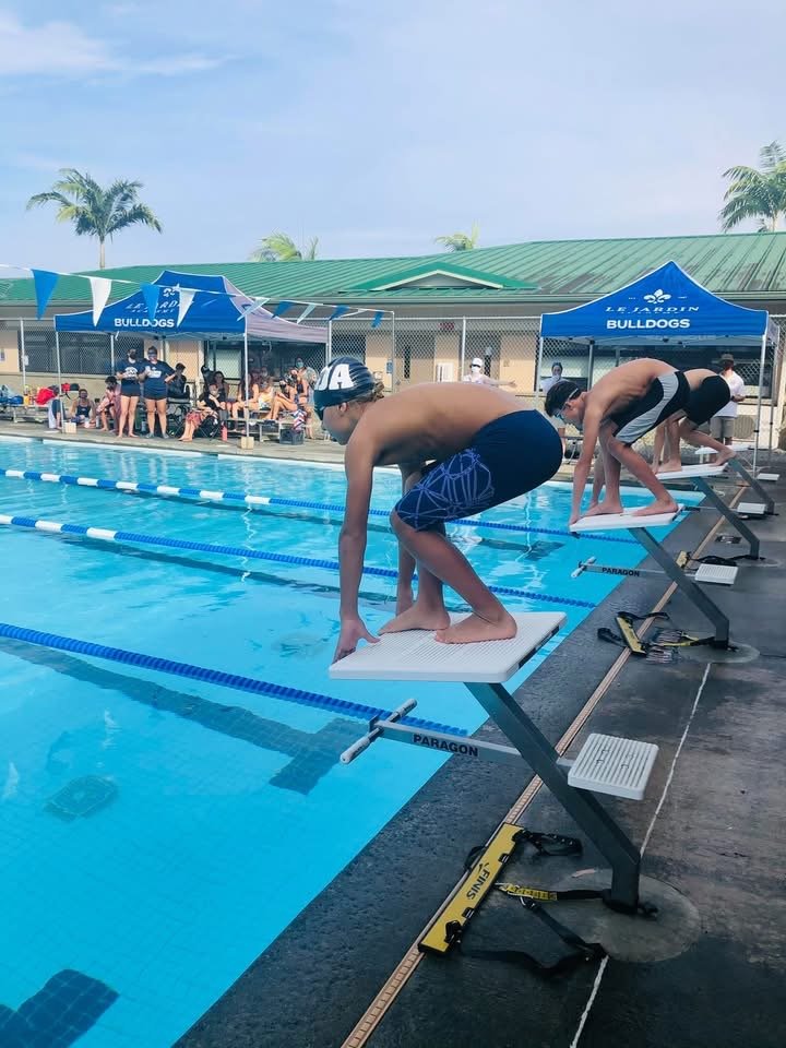 Two boys are about to dive into a swimming pool from starting blocks, with a group of people in the background under blue umbrellas with the words 'Le Jarden Bulldogs' on them.