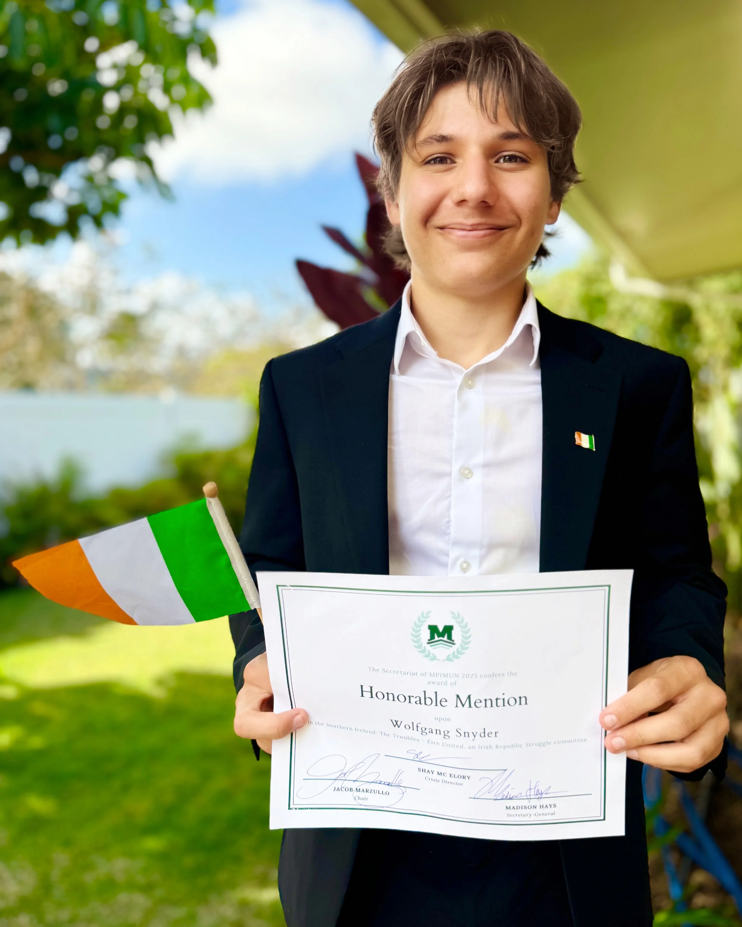 A young man in a suit holding an Irish flag and a certificate outdoors with green trees and blue sky in the background.