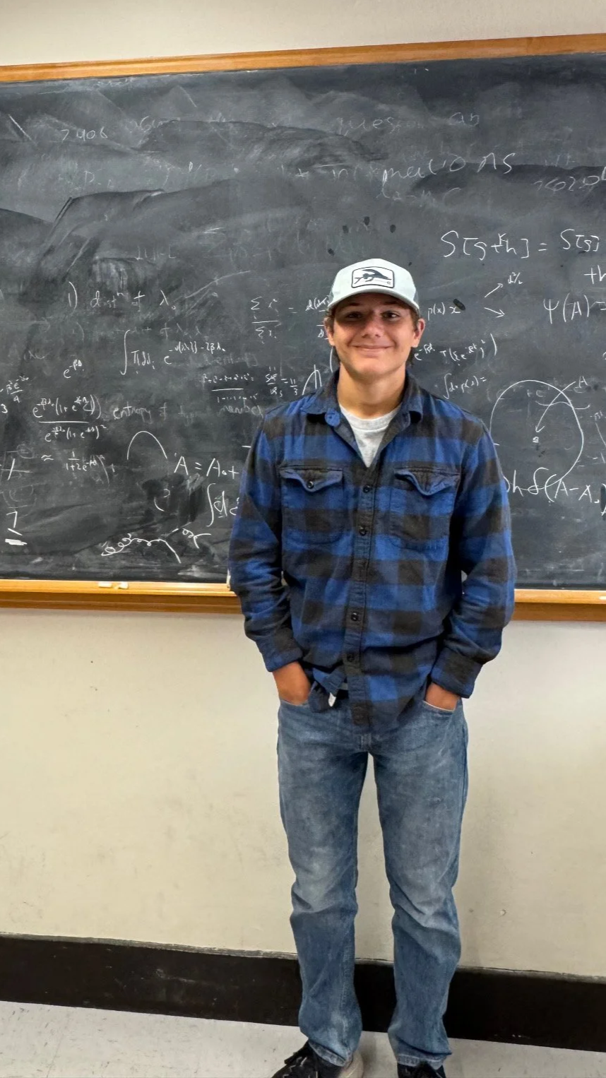 A young man standing in front of a blackboard filled with mathematical equations, wearing a white cap, blue and black checkered shirt, and jeans, smiling with hands in pockets.