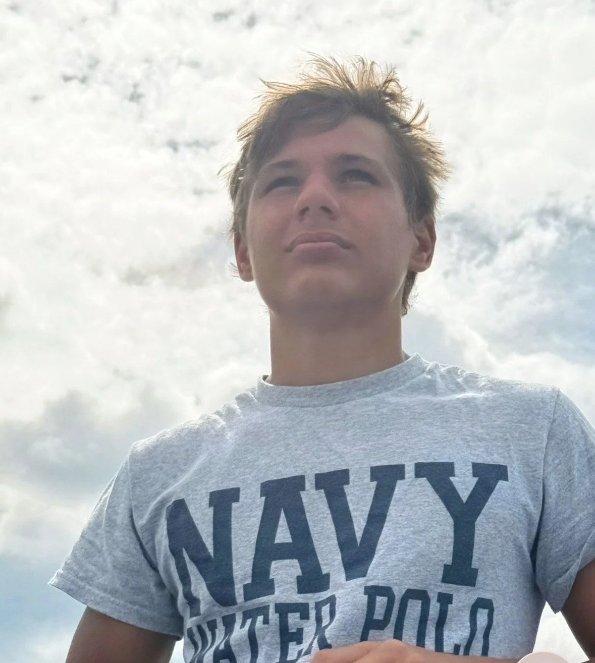 Young man with spiky hair wearing a gray t-shirt with 'NAVY' printed on it, standing outdoors against a cloudy sky.