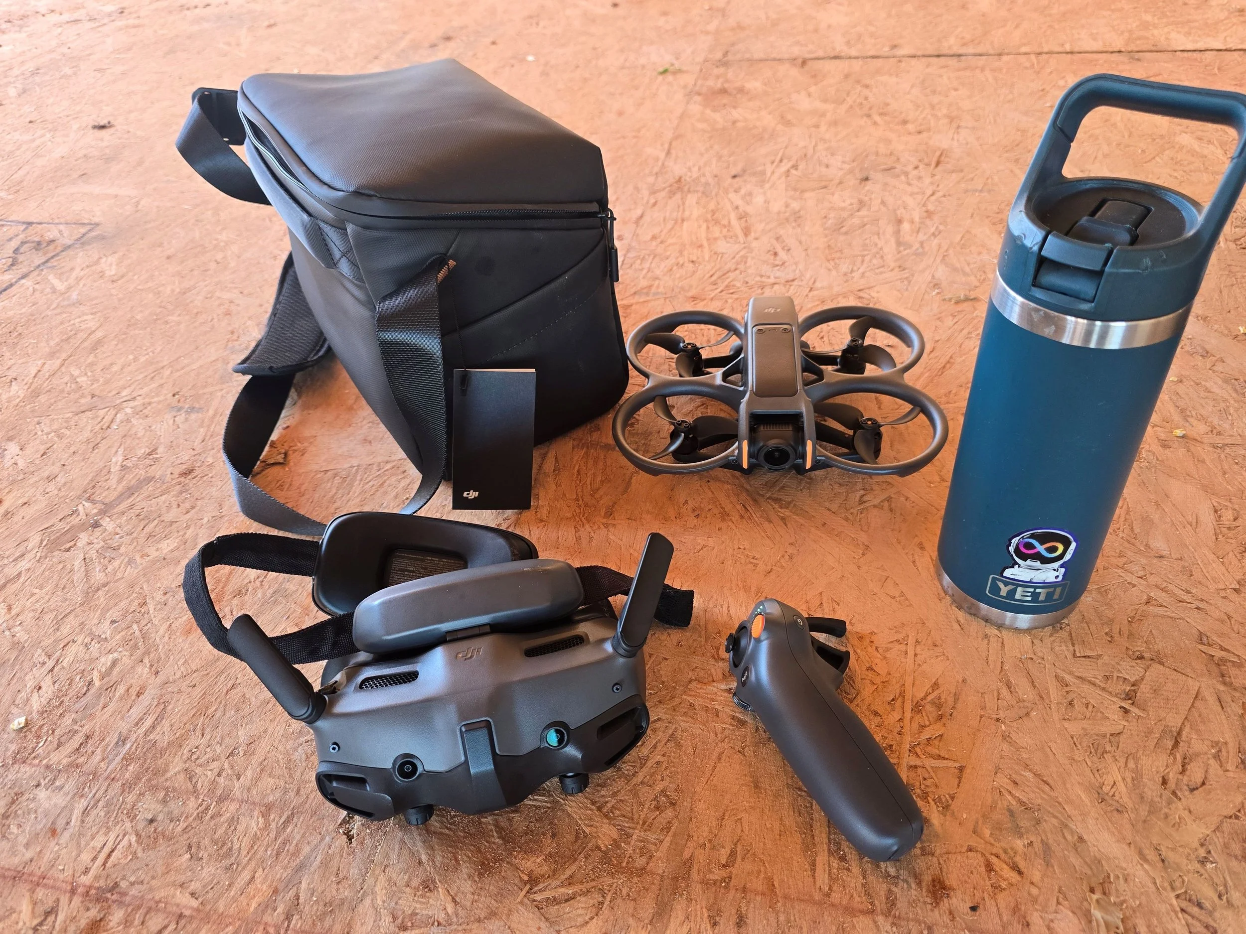 Black backpack, small drone with propellers, blue water bottle, and robot ride-on toy on a wooden floor.