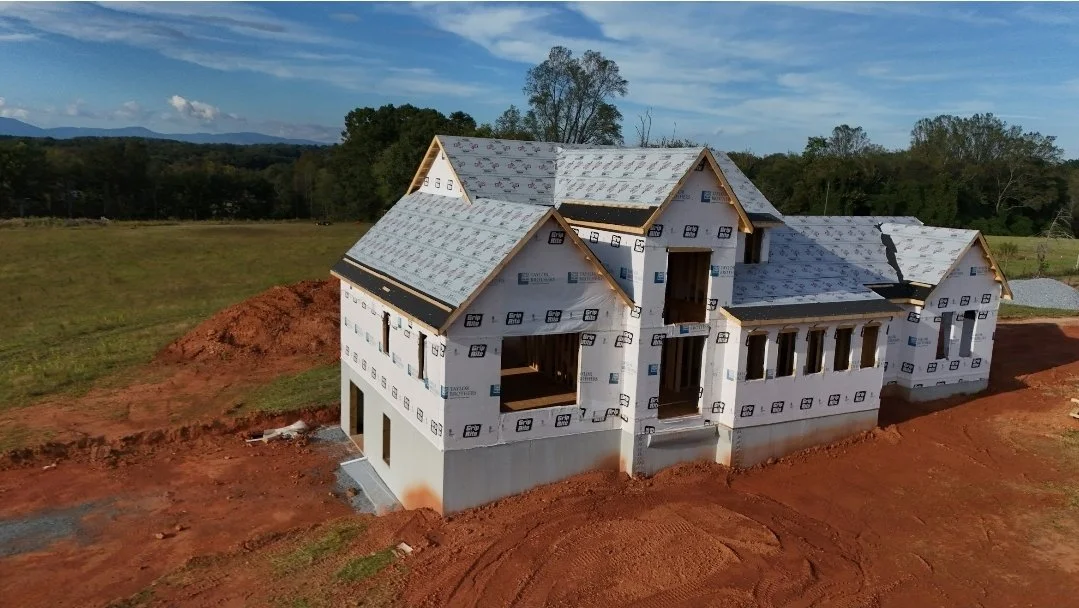 A house under construction with a partially finished roof and insulation on the walls, situated on a red dirt lot surrounded by grassy fields and trees.