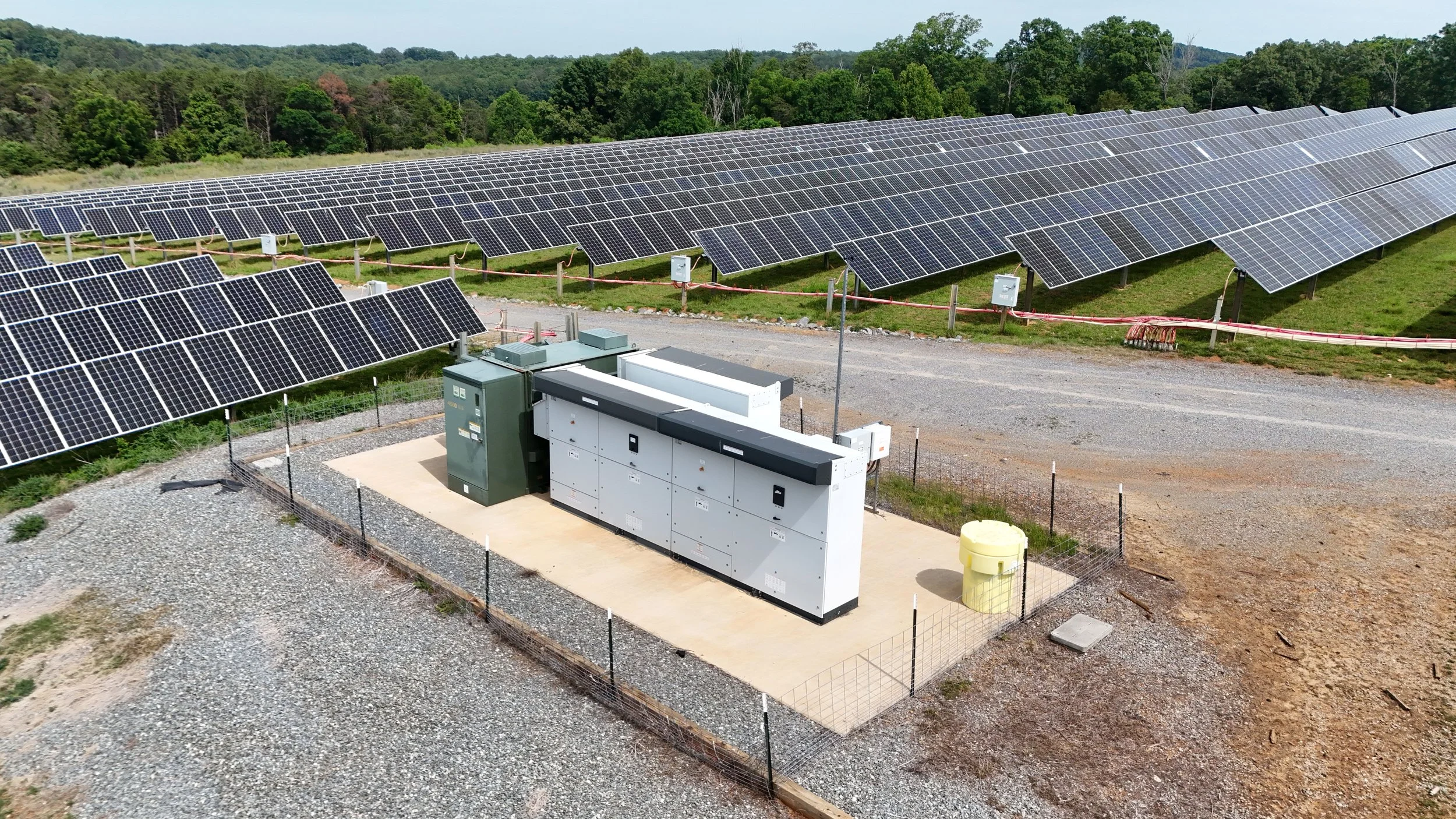 A large solar panel installation in a rural area with green trees and hills in the background, including electrical equipment and wiring in the foreground.