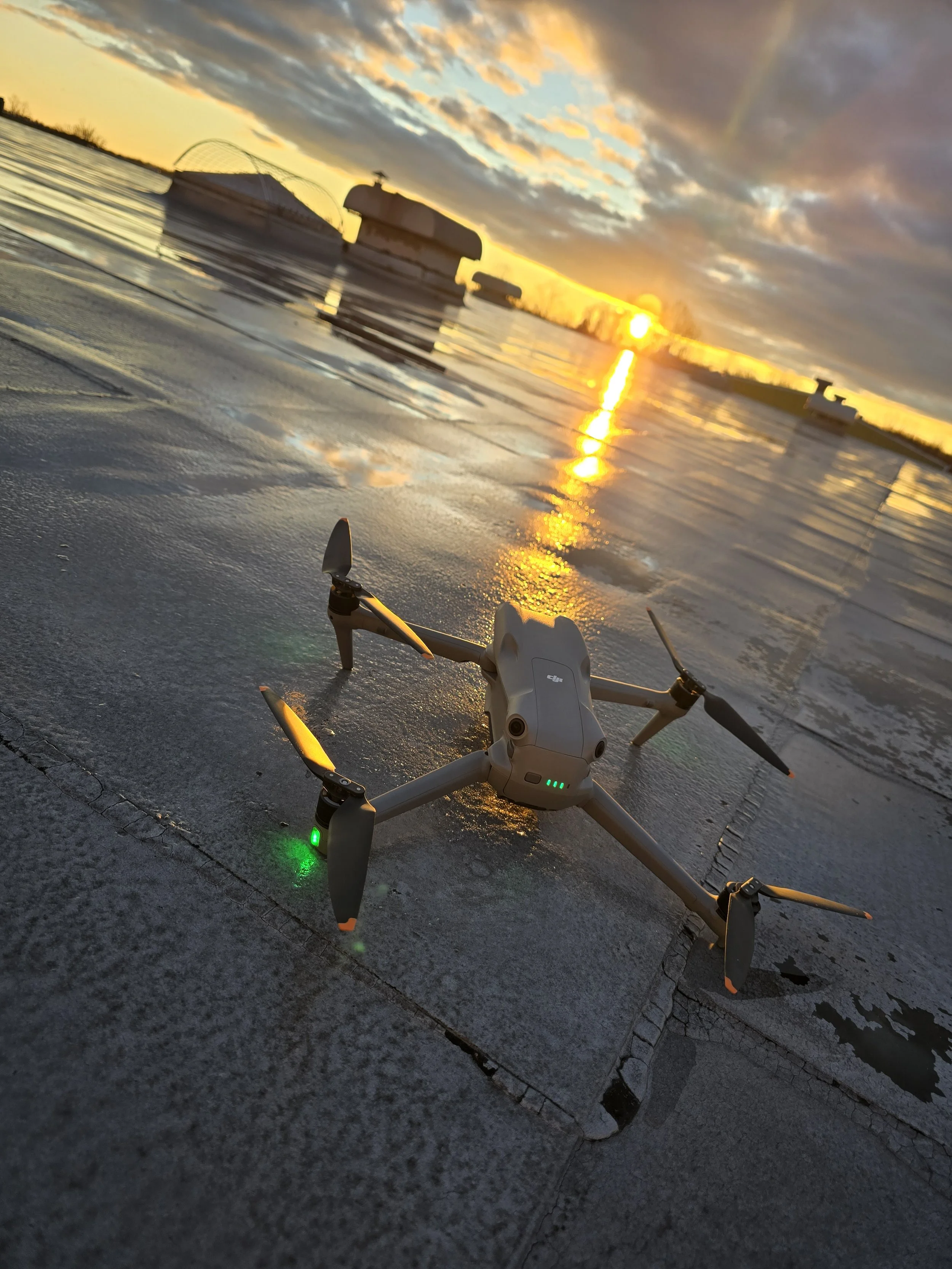 A drone on the ground near a body of water during sunset, with a cloudy sky in the background.