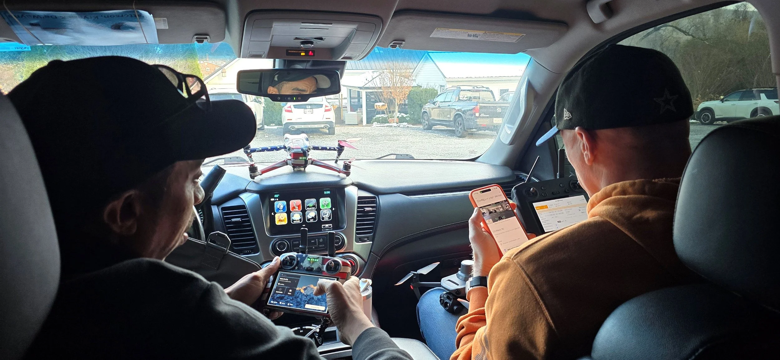 Two people sitting in the front seats of a vehicle, looking at their smartphones. The dashboard displays a GPS app, and there is a drone on the dashboard. The view outside shows a parking lot with several cars and a small building.