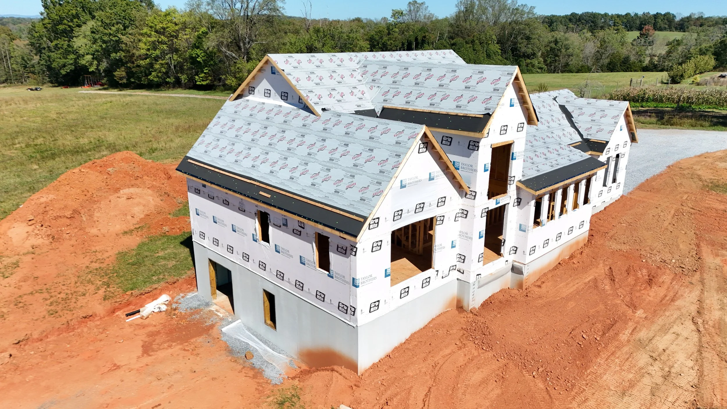 House under construction with a partially completed roof and white sheathing, surrounded by dirt and green fields, with trees in the background.