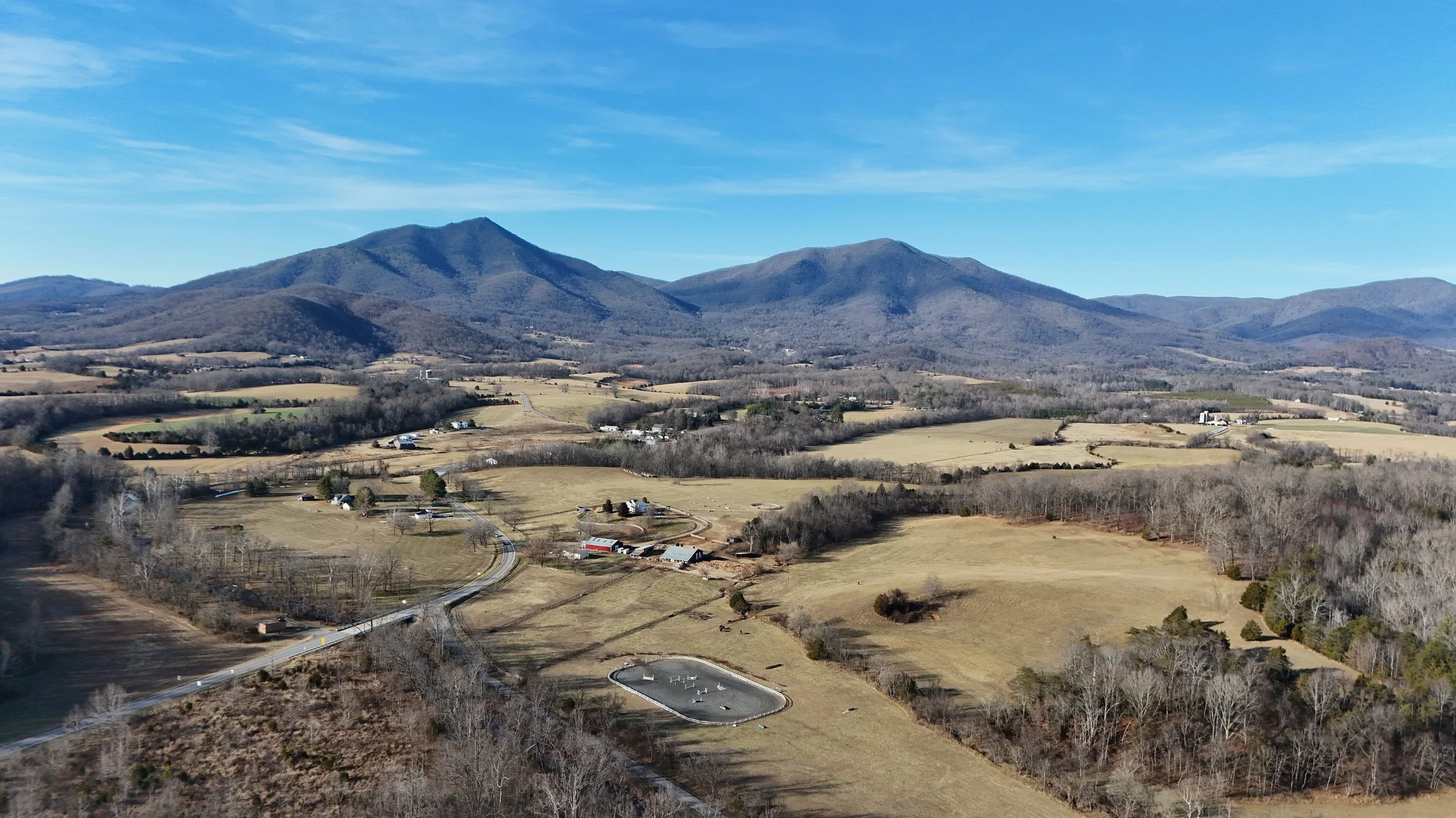 A scenic view of rolling hills and farmland with mountains in the background, a clear blue sky, and scattered trees and buildings.
