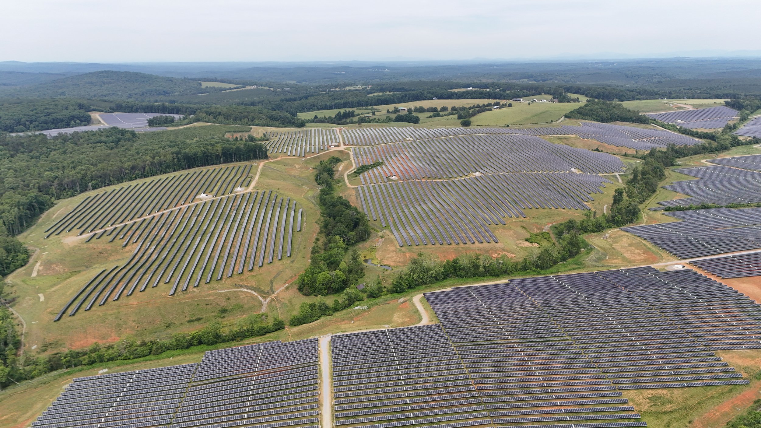 Aerial view of a large solar farm with numerous solar panels arranged in rows across hills and fields surrounded by trees and distant mountains.