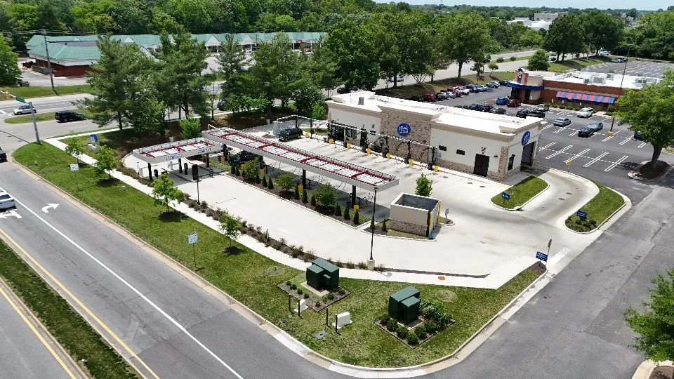 A drive-thru area with covered lanes, a small building, and parking lot, surrounded by trees, in a commercial area.