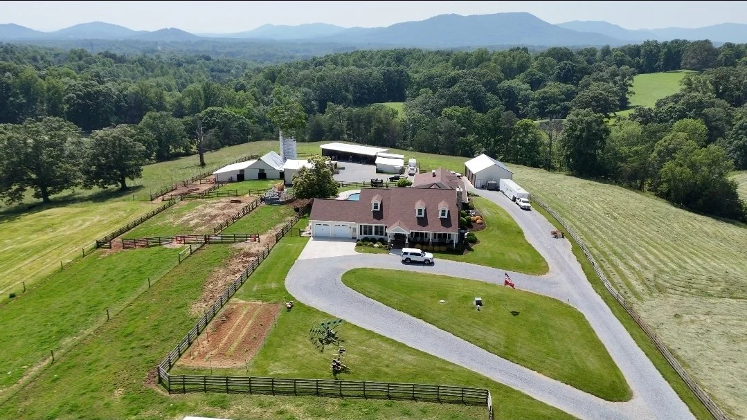Aerial view of a rural property with a house, multiple outbuildings, fenced fields, and surrounding green landscape with hills in the background.