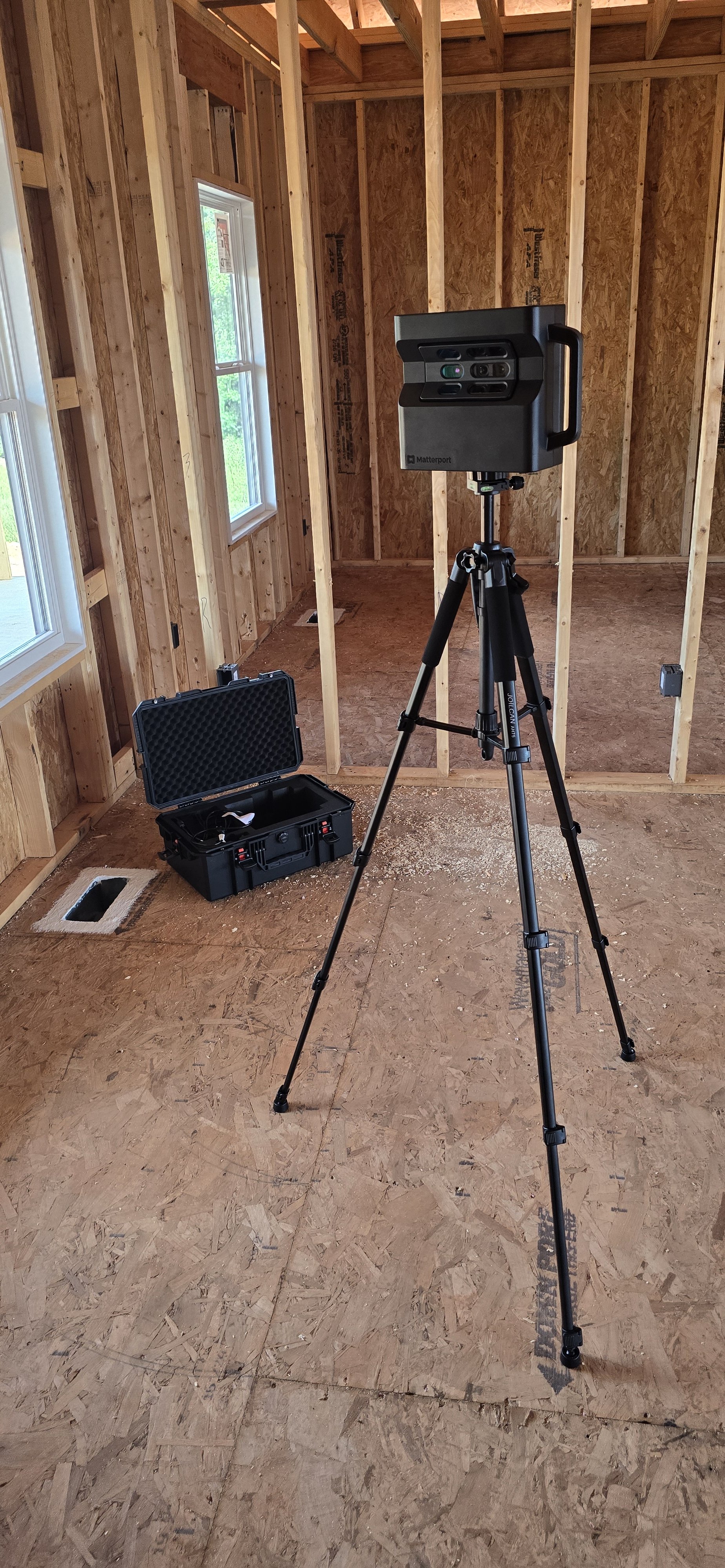 Construction site interior with wooden framing, two windows, a tripod with a 3D laser scanner, and a black hard case on the floor.