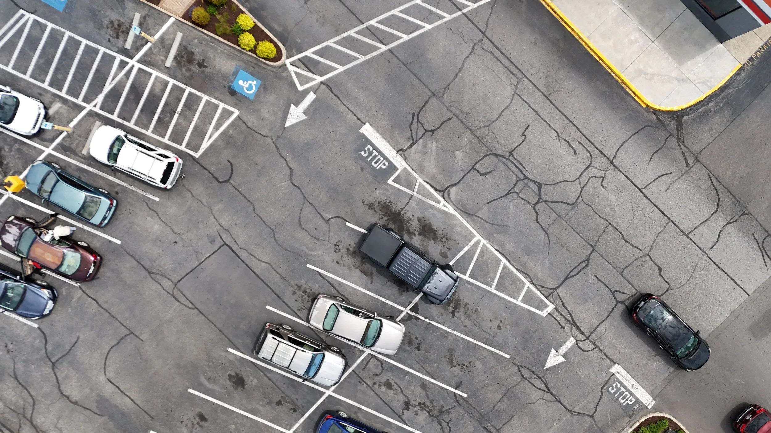 An aerial view of a parking lot shows several parked cars and empty spaces, including a designated handicapped parking space and a space with a small black truck. The asphalt has visible cracks, and there are small landscaped areas with green bushes.