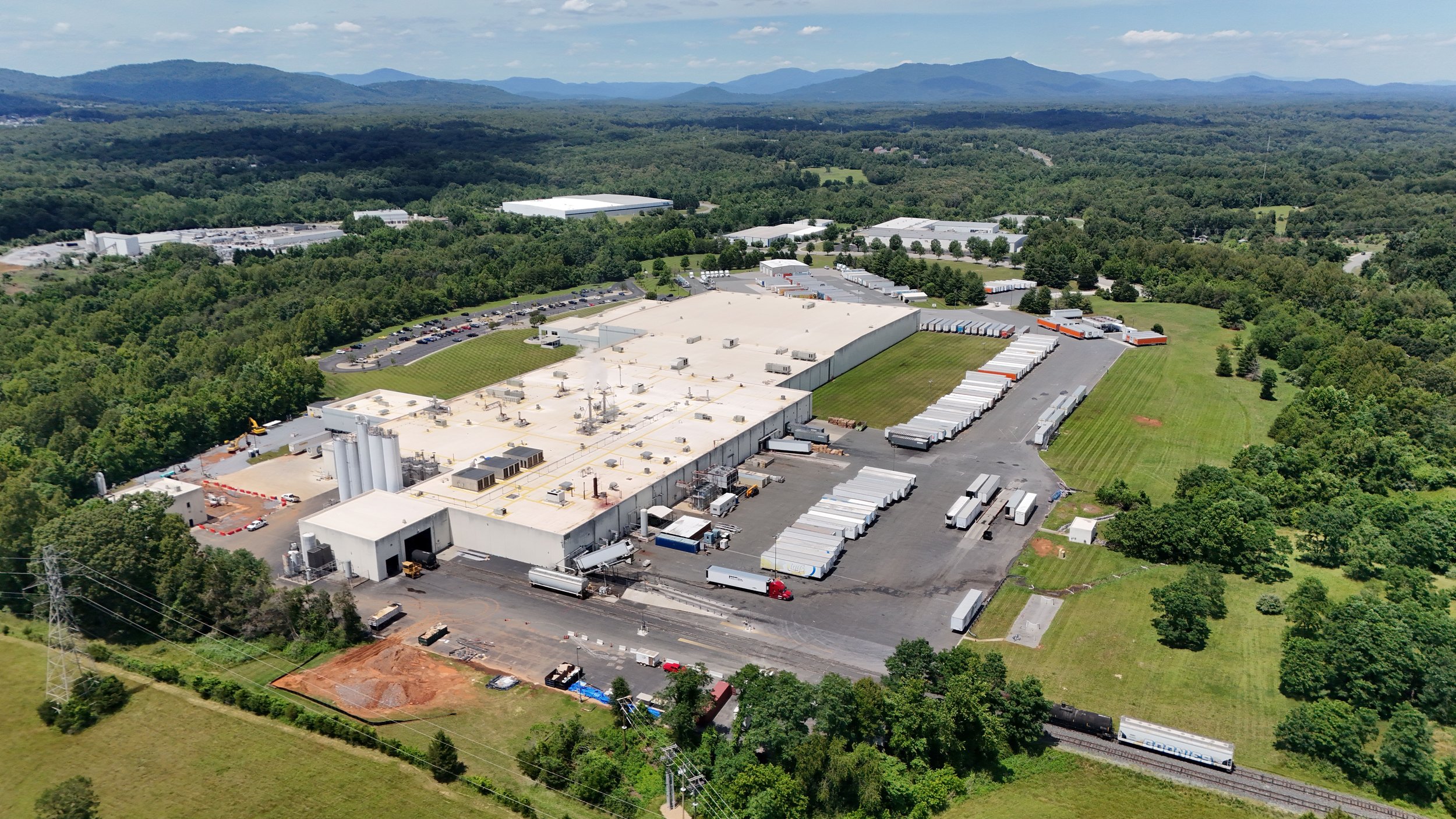 Aerial view of a large industrial complex surrounded by green trees and rolling hills in the background.