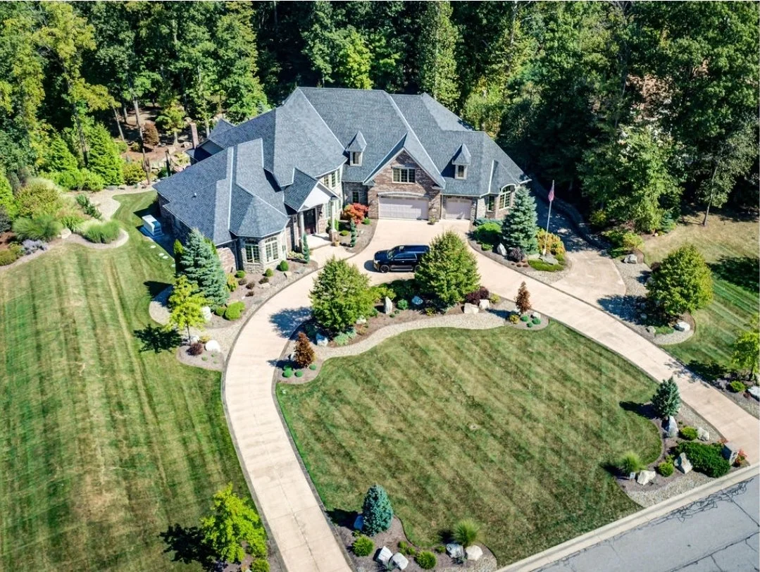 A large, upscale house with a gray roof, surrounded by a well-maintained lawn and landscaped yard, with trees and bushes, and a curved driveway leading to the garage.