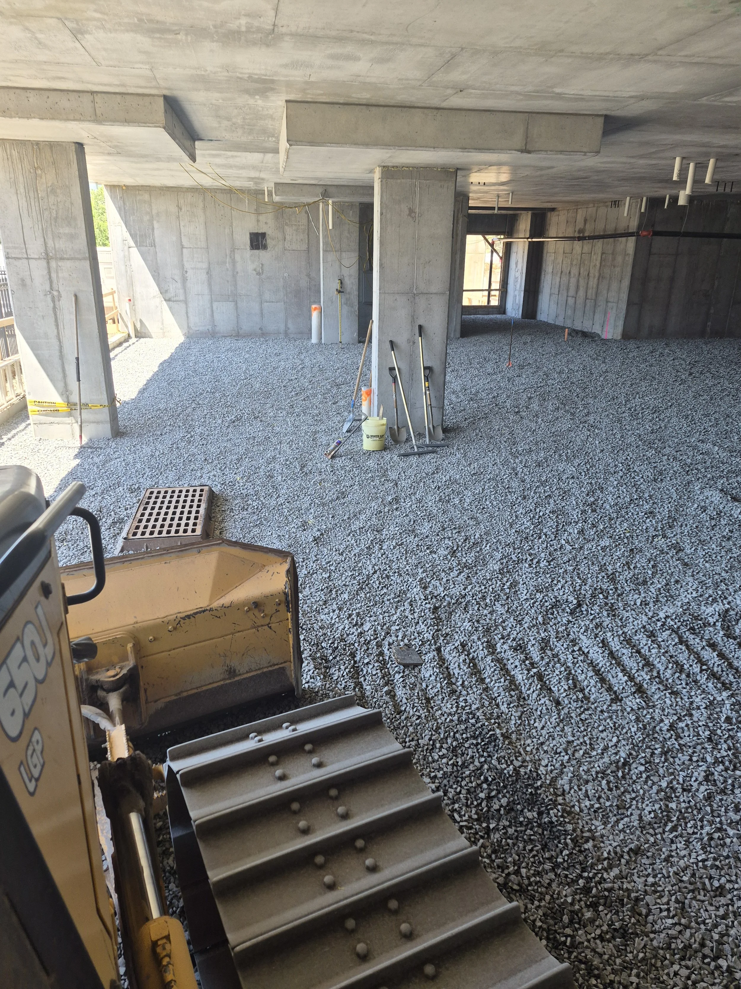 Construction site with gravel-covered ground, concrete walls and ceiling, tools and equipment including a shovel, a bucket, and a mini excavator, with pipes and unfinished electrical wiring visible.