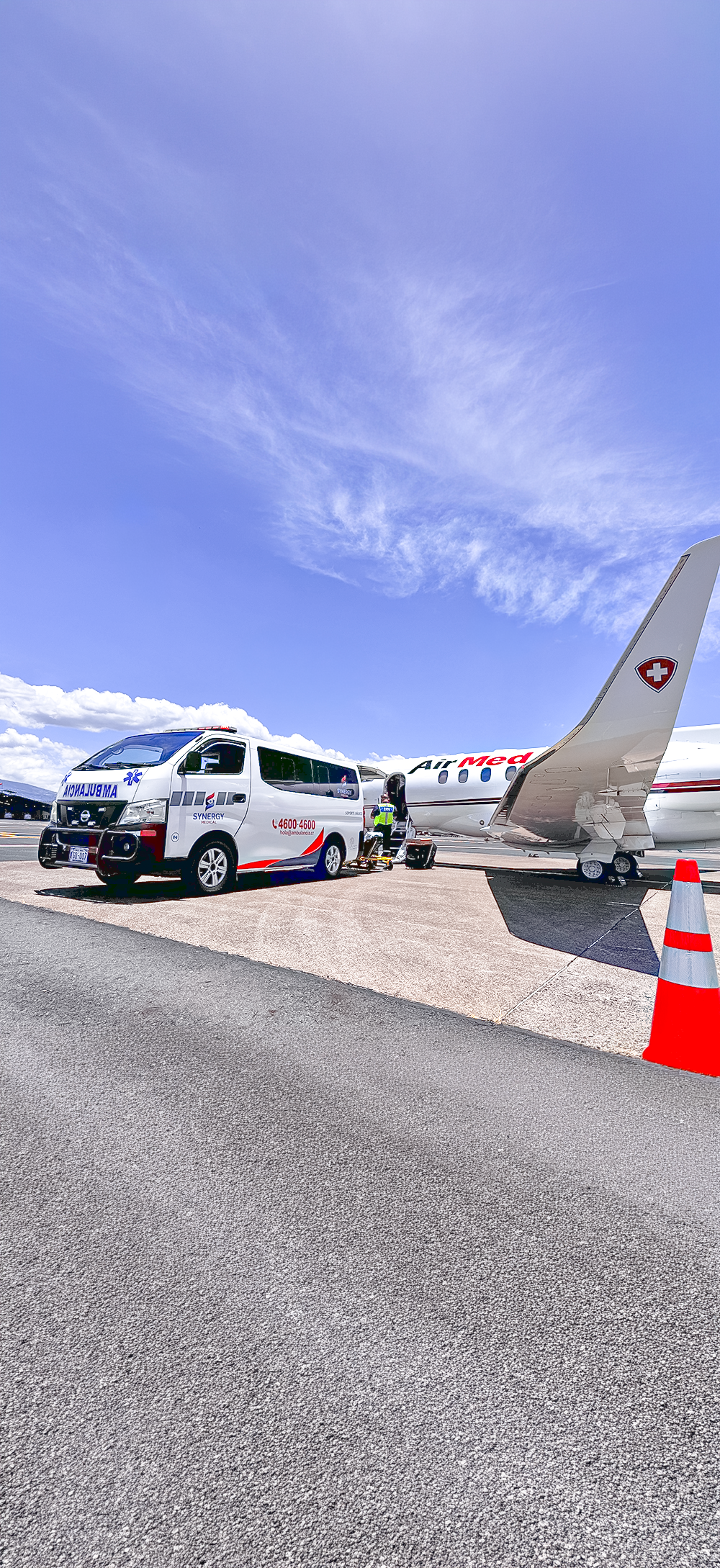 Emergency medical team preparing an ambulance next to a private jet on the tarmac under a cloudy blue sky.