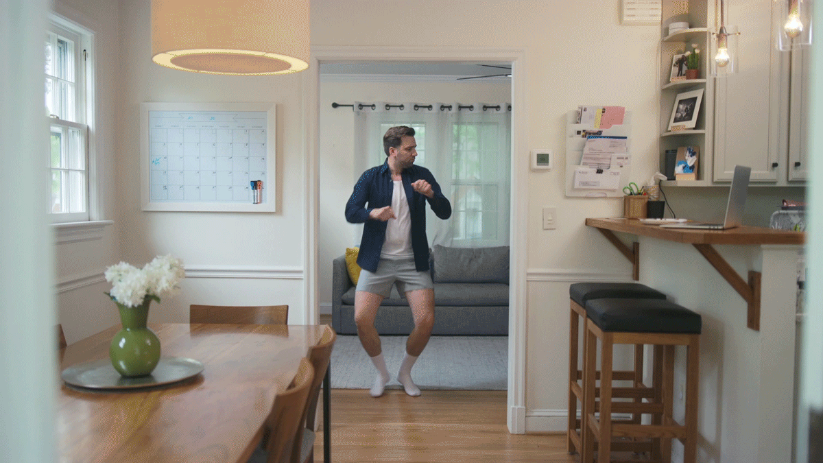 A man in boxers and a shirt dancing in a kitchen, with a living room visible in the background.
