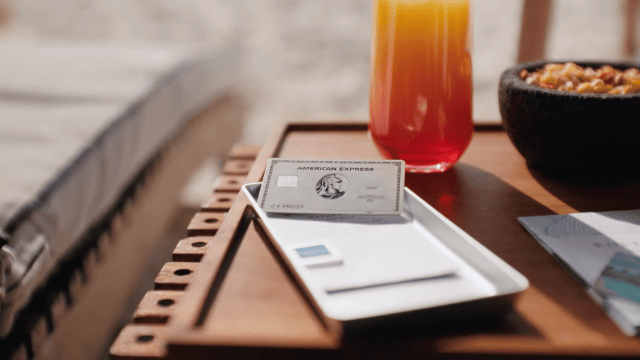 A tray with a credit card, a paper receipt, a glass of orange-colored beverage, a bowl of snack mix, and some papers on a wooden table.