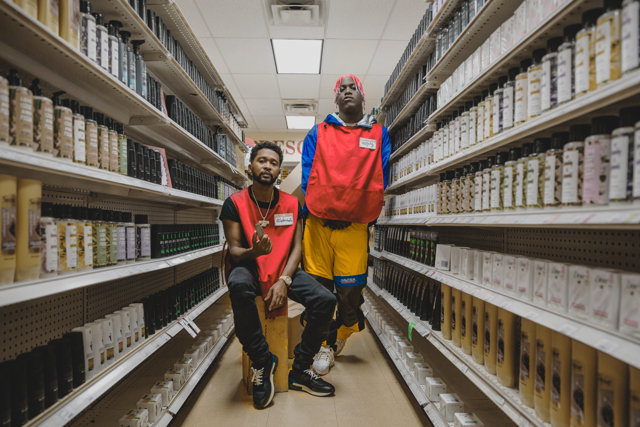 Two young men in colorful streetwear pose in the dairy aisle of a grocery store, surrounded by shelves filled with bottles of lotion or shampoo.