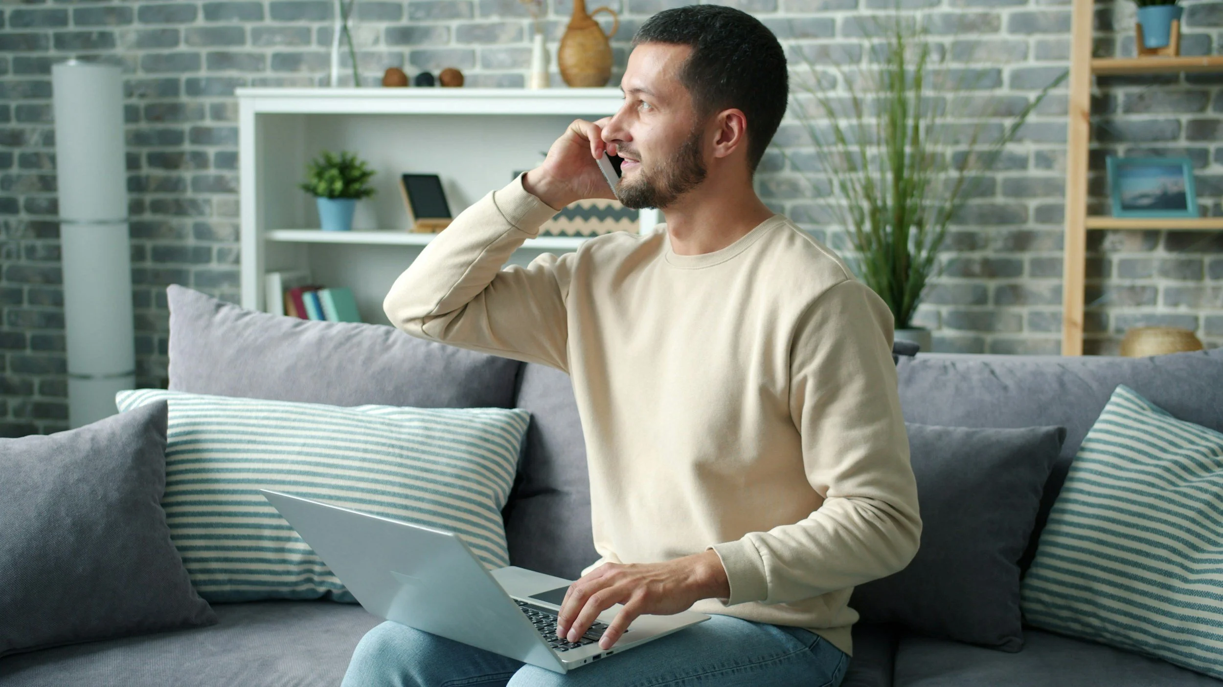 A man with short dark hair and a beard, wearing a beige long-sleeve shirt, is sitting on a gray couch with striped and solid pillows. He is holding a cellphone to his ear with his right hand and using a laptop with his left hand. The background shows a brick wall, a white bookshelf with a green potted plant, and decorative items including vases and framed pictures.