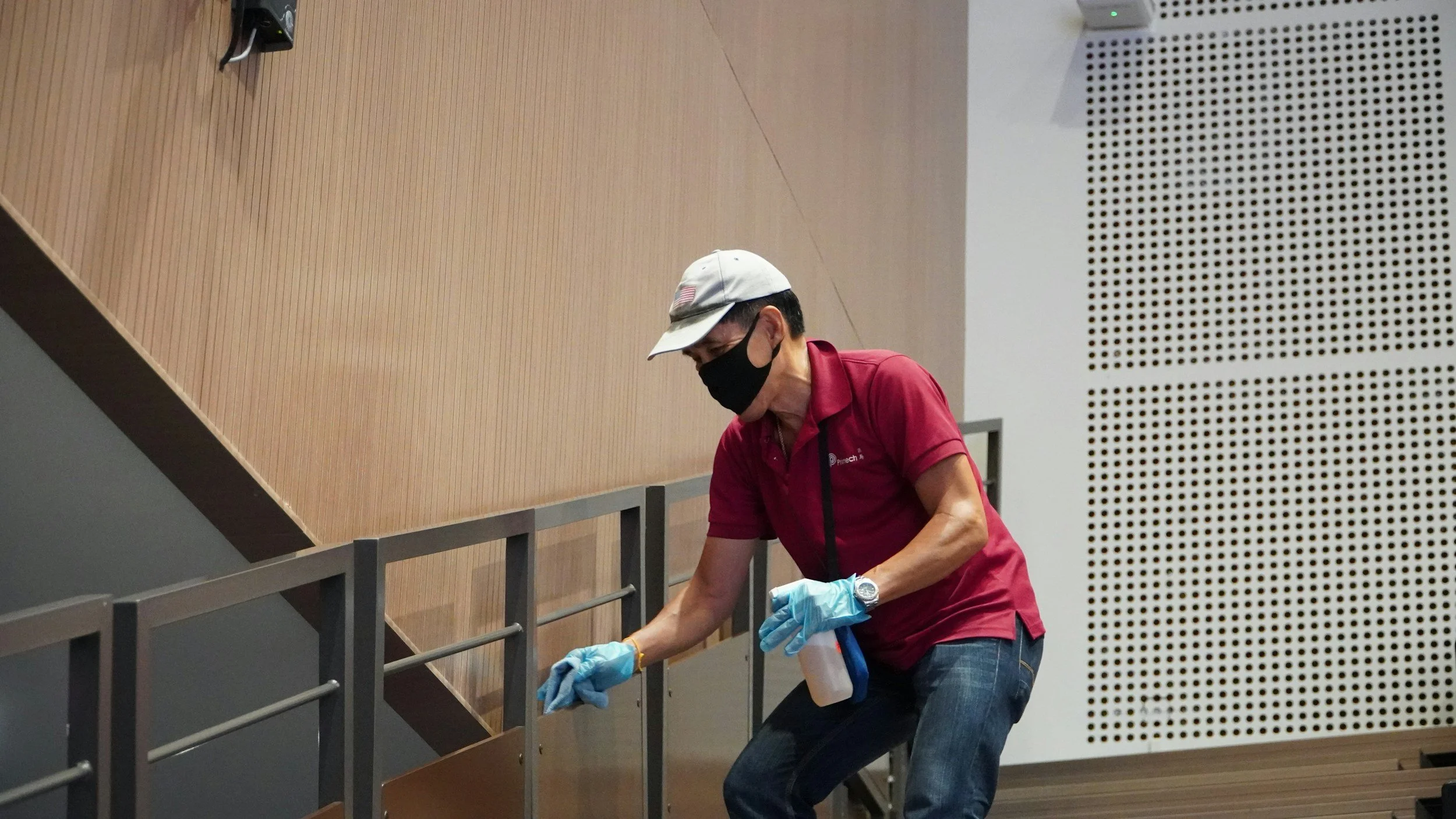 A man wearing a beige cap, black face mask, red shirt, and blue gloves cleaning or sterilizing a railing inside a room with wood-paneled wall and decorated white wall with small perforations.