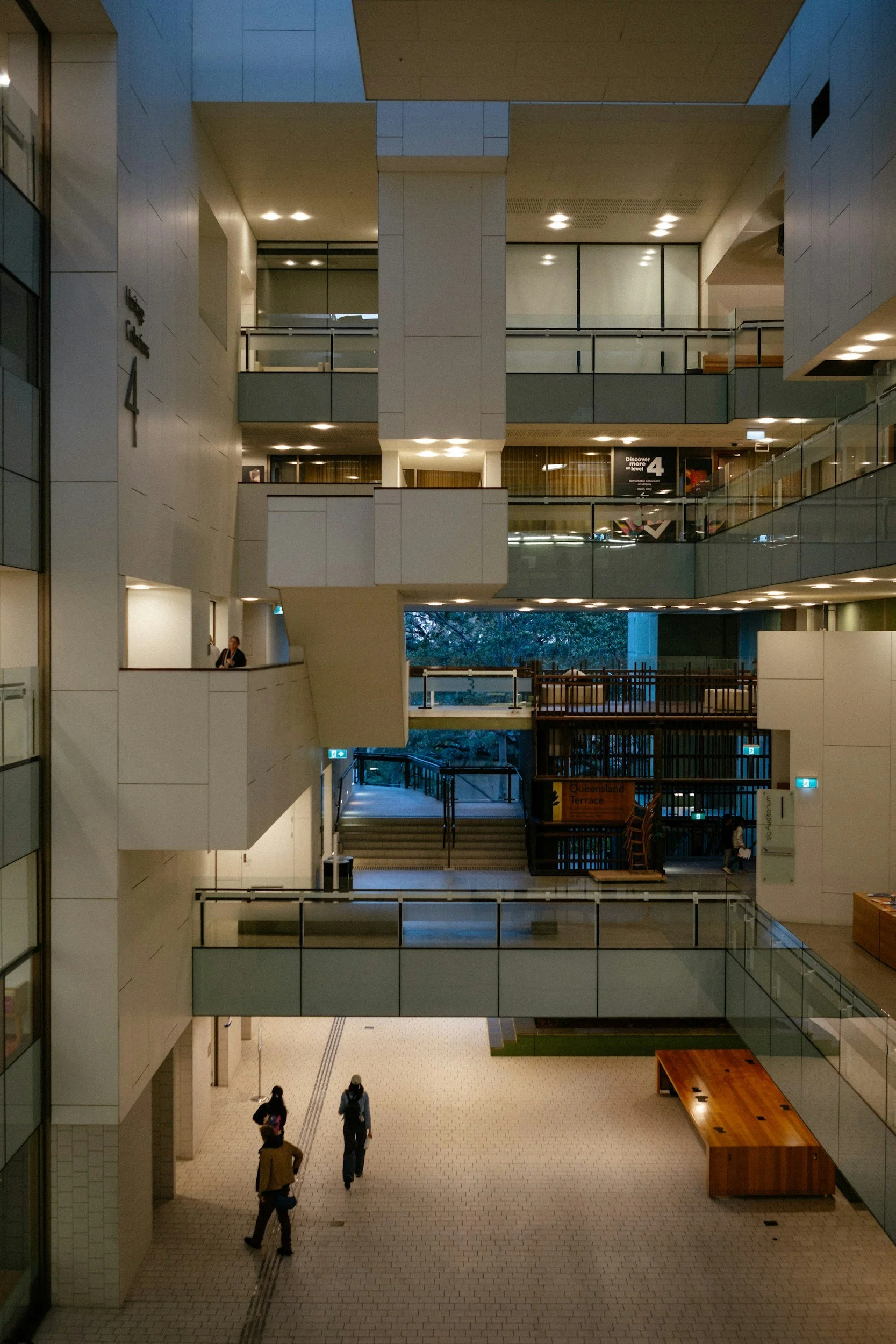 Interior of a modern multi-level building with glass and concrete architecture. Several people are walking on the tiled floor, some carrying bags. Bright lighting illuminates the open space with multiple balconies and railings visible on upper floors. There are signs indicating levels and directions, including a sign for Queensland Terrace.