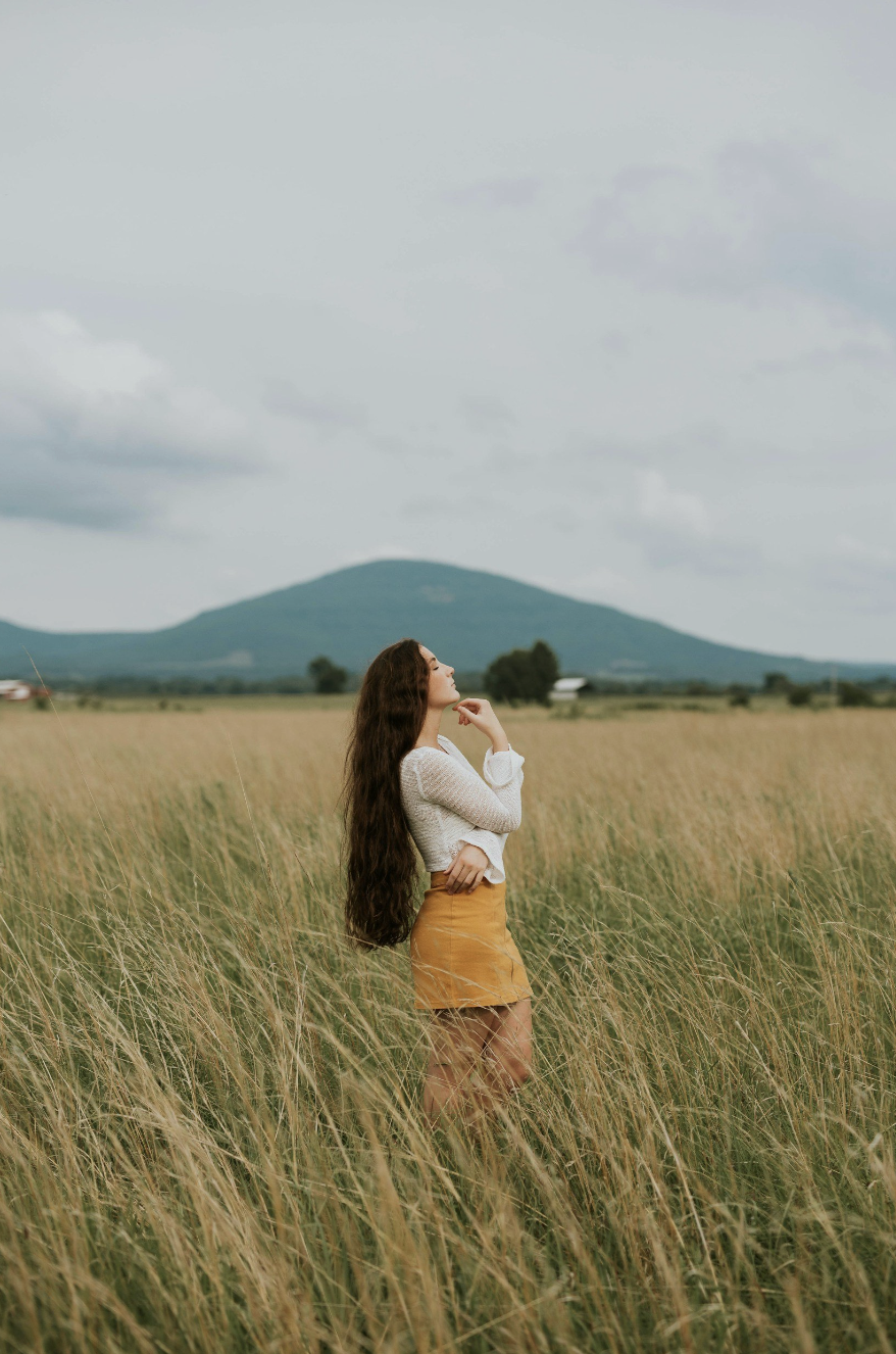 A woman with long brown hair standing in a grassy field, looking up towards the sky with a mountain in the background.