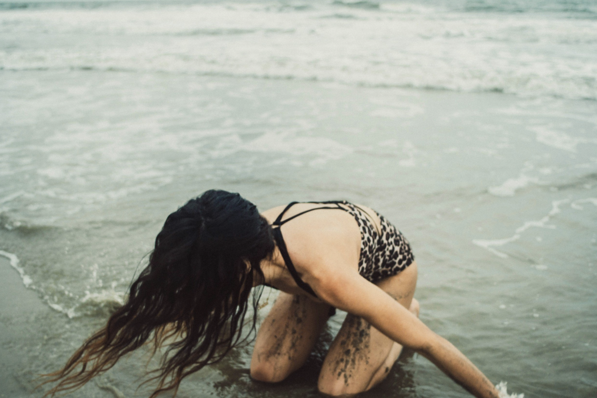 A woman with long dark hair wearing a leopard print swimsuit is kneeling on the wet sand at the beach with the ocean waves in the background.
