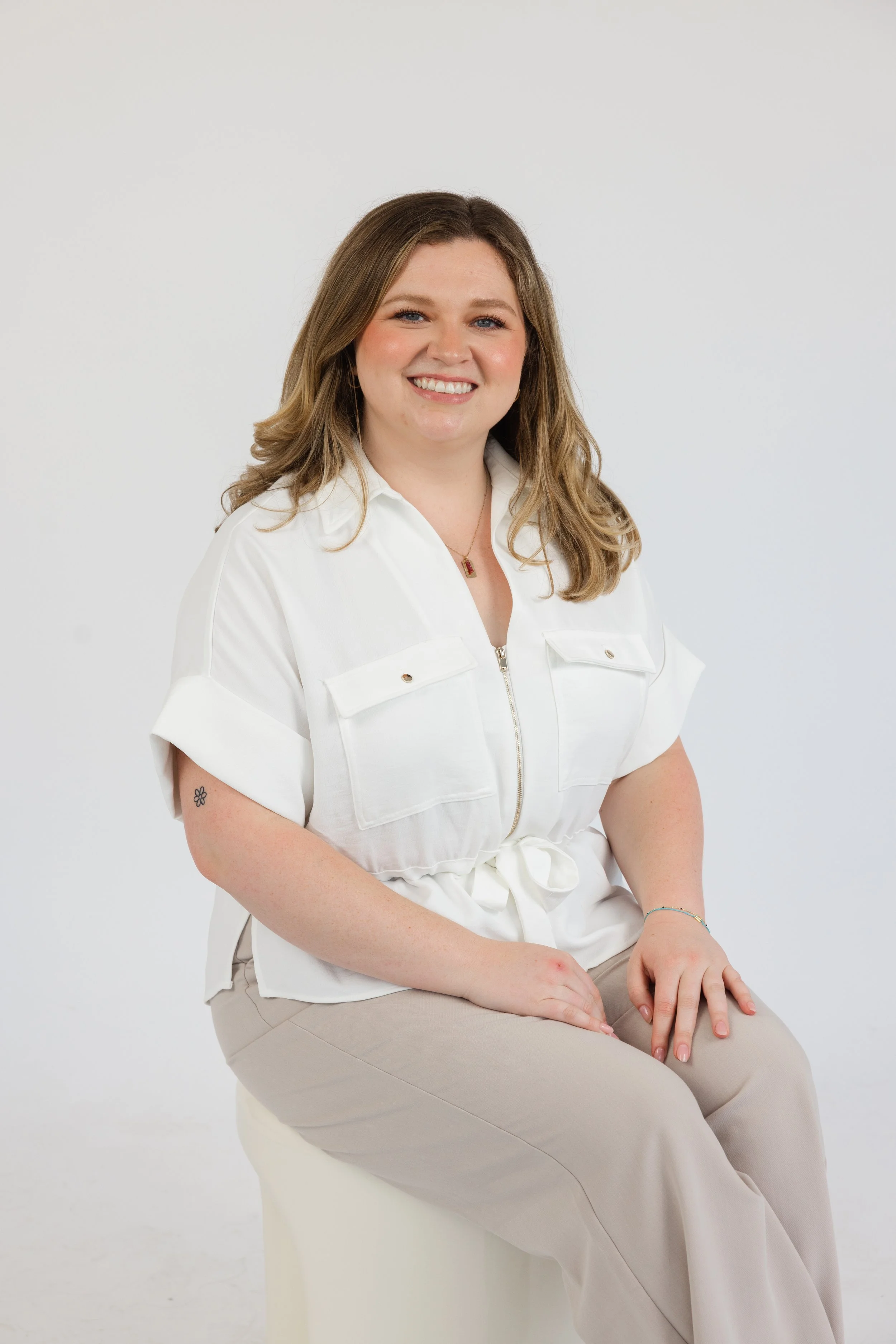 A woman with shoulder-length, wavy light brown hair, smiling with her teeth showing, wearing a white short-sleeved shirt with a front zipper and pockets, beige pants, and sitting on a white chair in front of a plain white background.