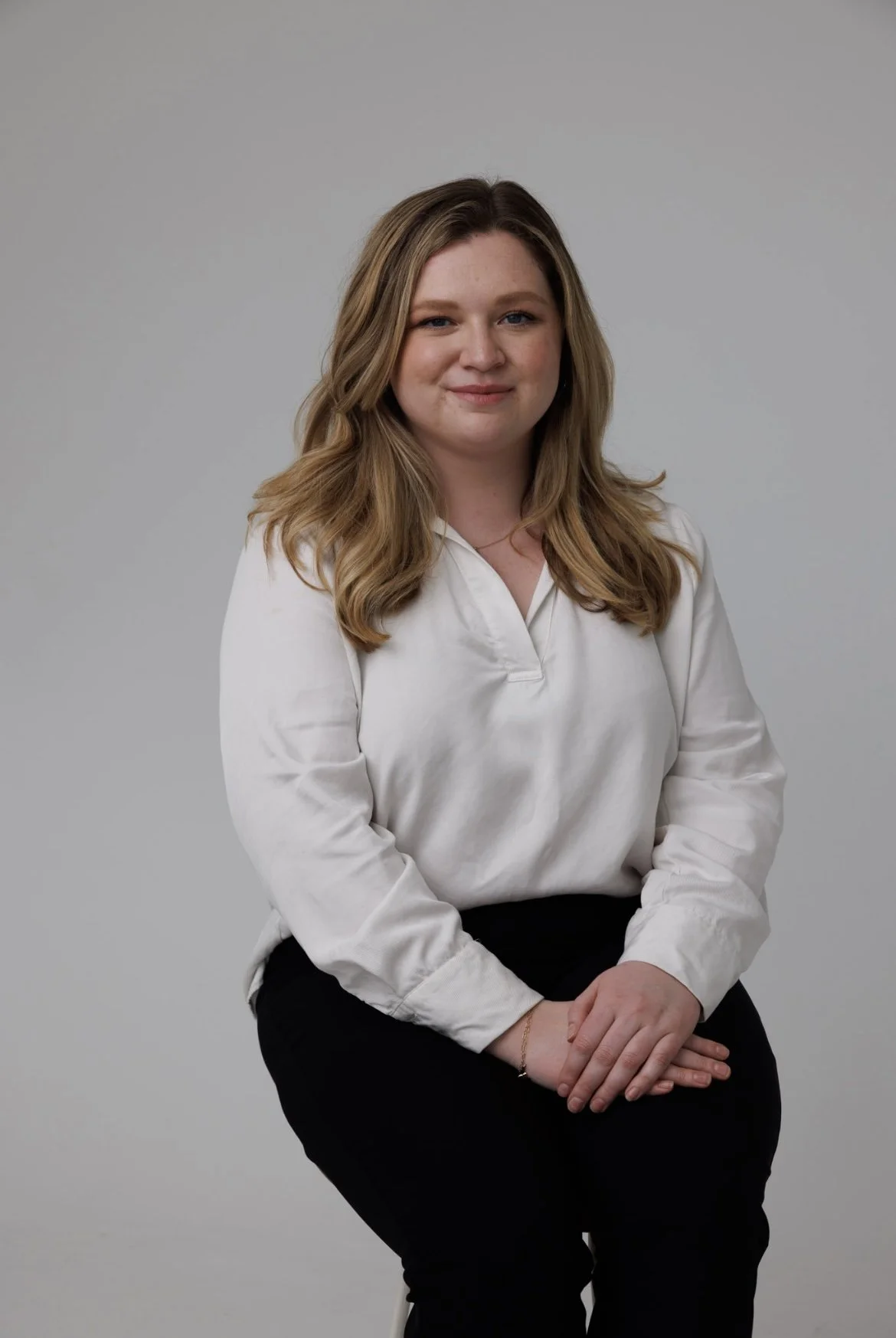 A woman with long, wavy, light brown hair wearing a white blouse and black pants, sitting against a plain gray background.