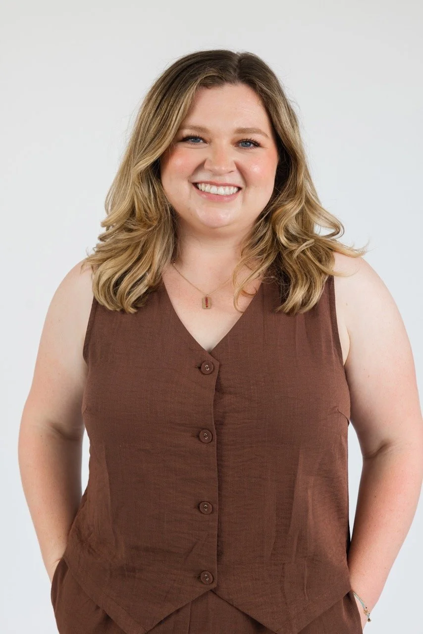 A smiling woman with shoulder-length wavy blonde hair in a sleeveless brown button-up top. She is standing against a plain white background.