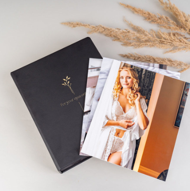 A black photo album with gold lettering and a stack of photos, including a woman with curly blonde hair in a white lace robe, on a white surface with dried beige ornamental grass.