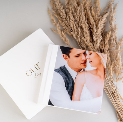A wedding album titled 'Our' with a photo of a bride and groom on the cover, placed next to dried pampas grass on a white surface.
