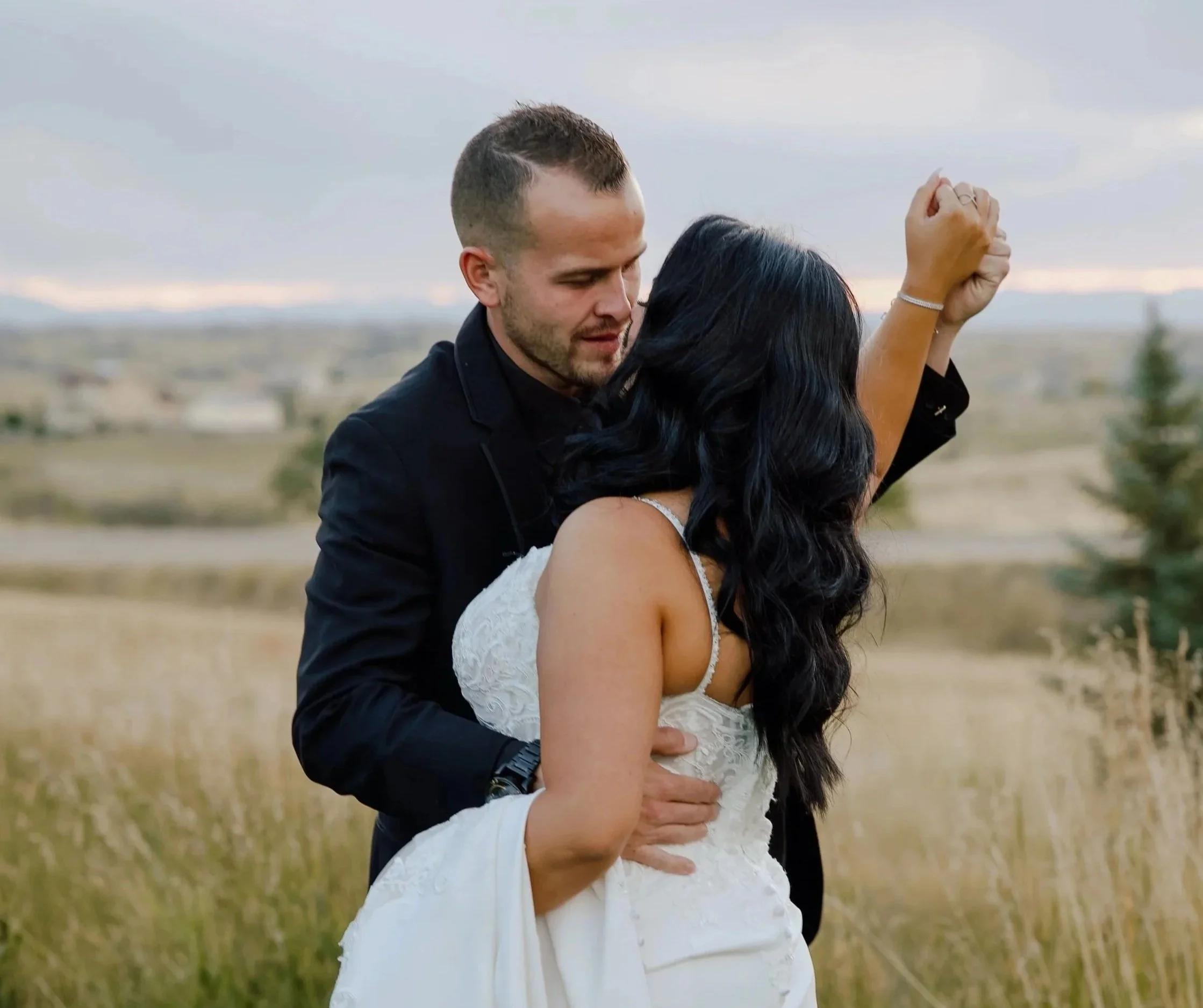 A couple is embracing outdoors in a field, with the man with short brown hair and a beard holding the woman from behind, both with their eyes closed, during sunset or late afternoon.