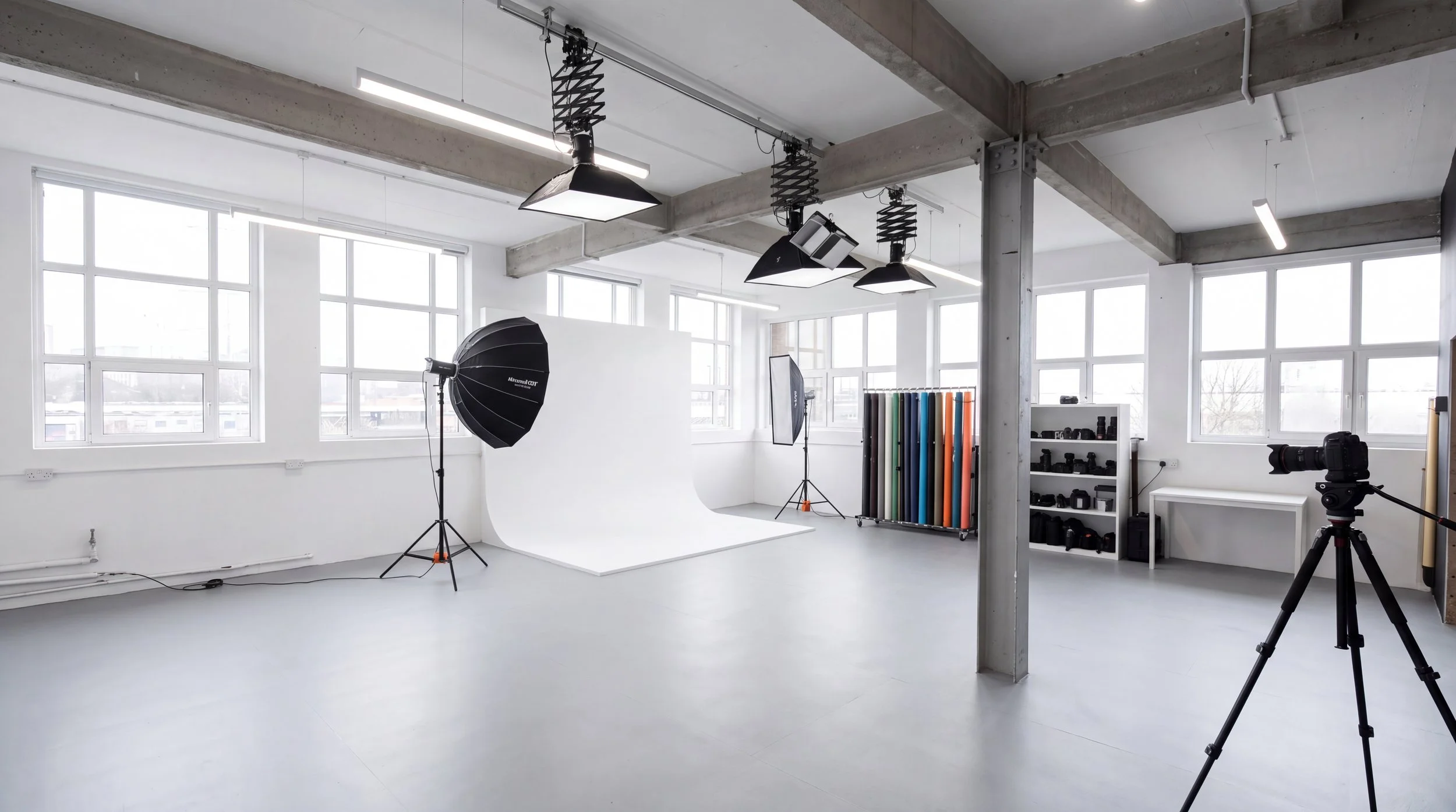 Photography studio with large windows, white backdrop, studio lighting equipment, and camera on tripod.