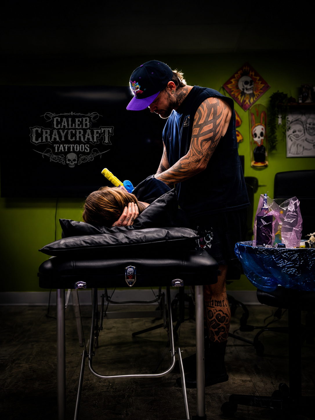 Tattoo artist tattooing a customer at a tattoo shop with a green wall decorated with skull artwork.