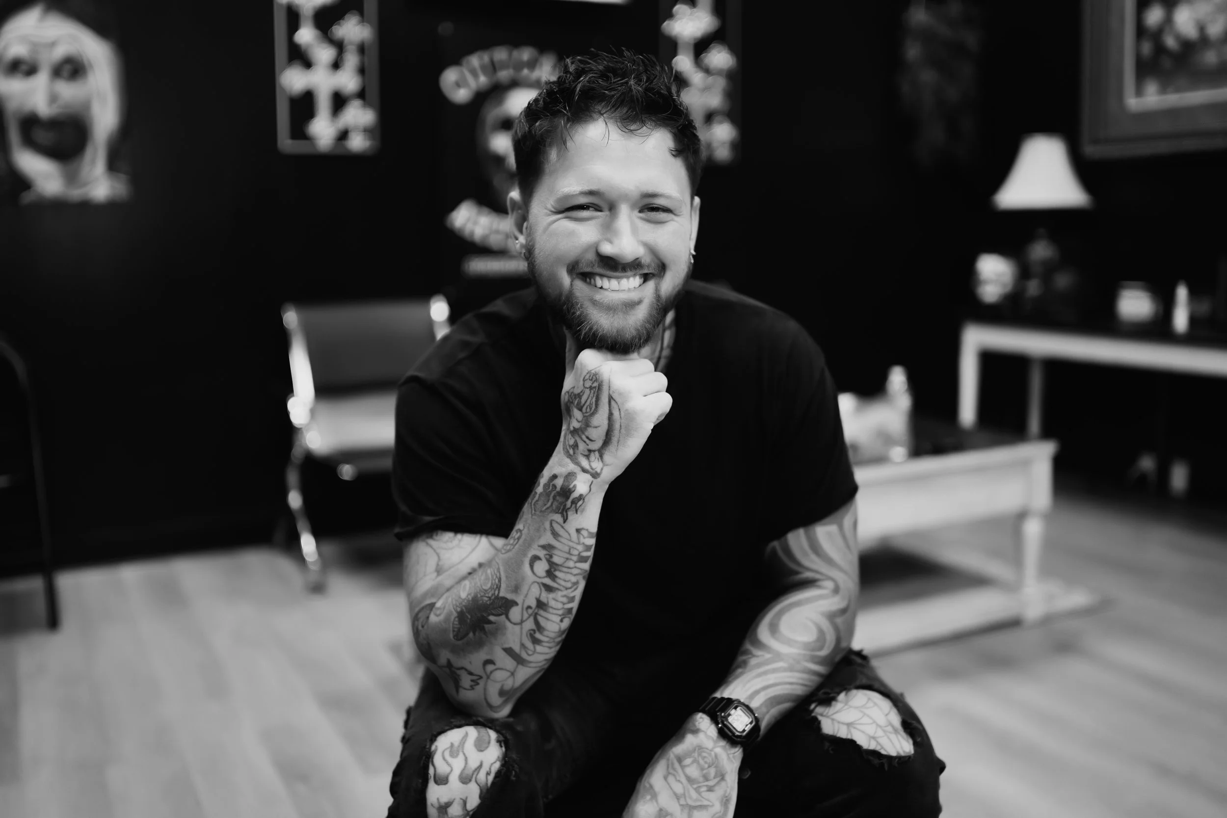 A smiling young man with tattoos on his arms, sitting on the floor in a tattoo studio, with tattoos on the walls and tattoos supplies in the background.