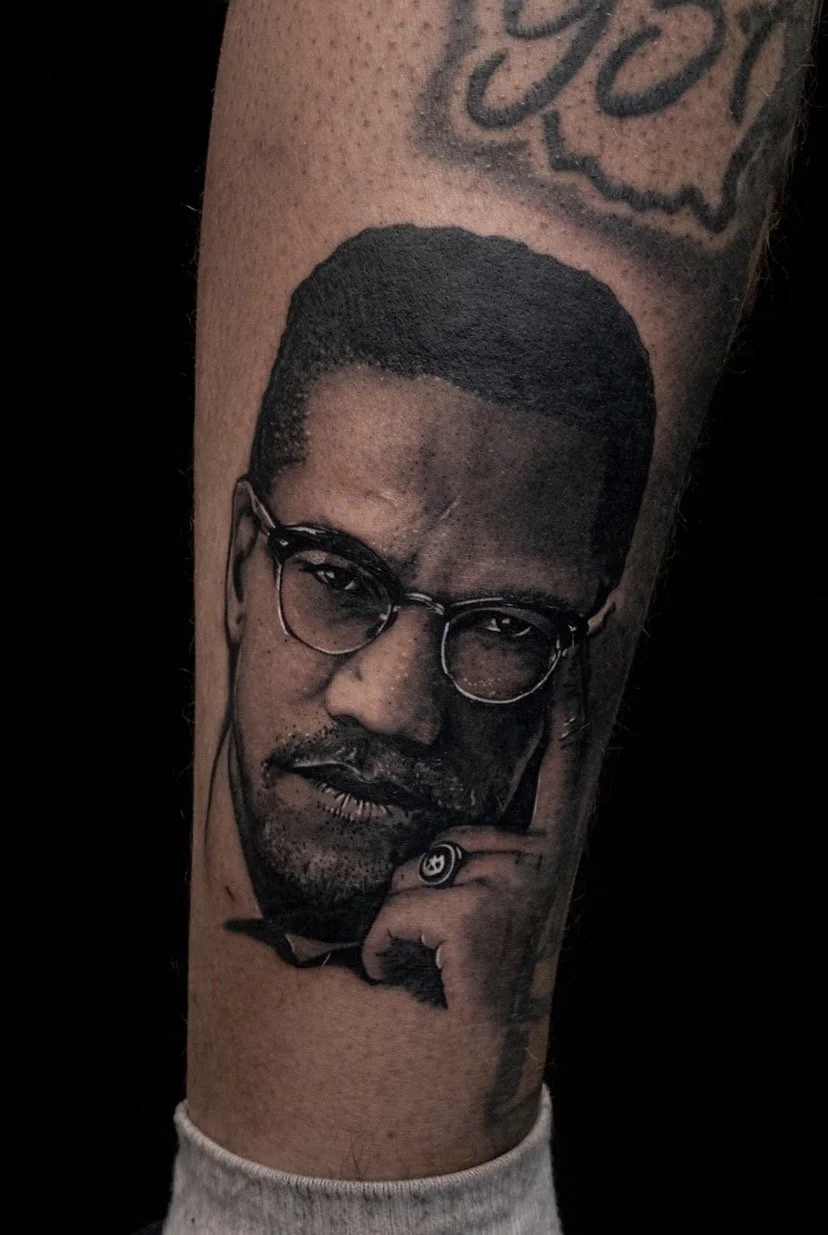 Black and gray realistic portrait tattoo of a man with glasses, a beard, and mustache, holding his chin with his hand, wearing a ring, with another tattoo partially visible above.