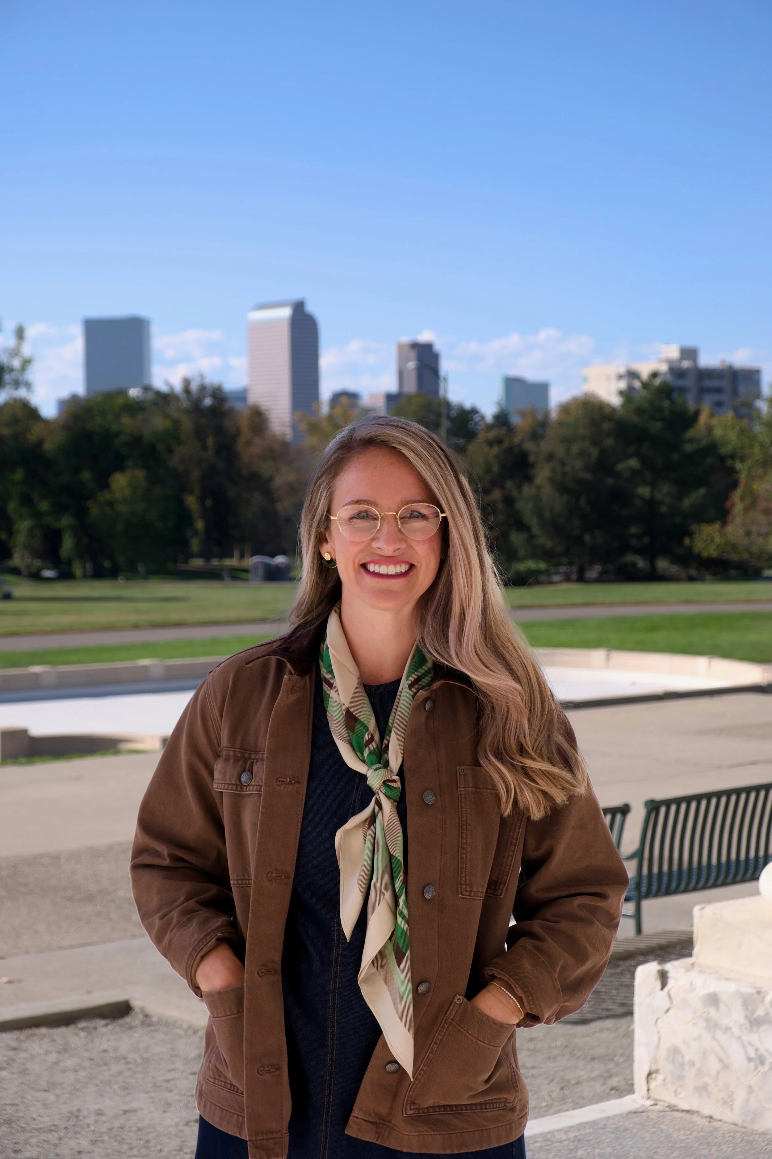 Smiling woman wearing glasses, a brown jacket, and patterned scarf in a city park with skyscrapers in the background