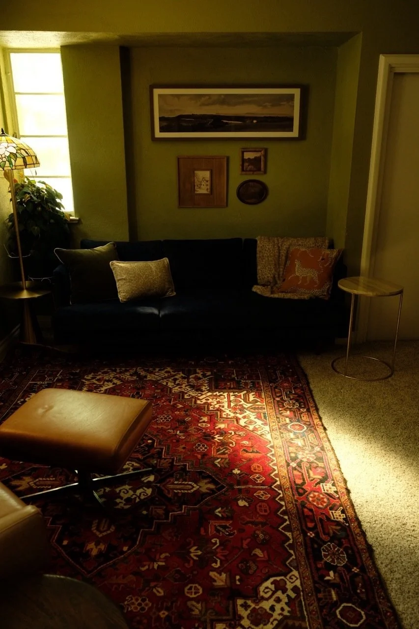 Cozy living room with a dark sofa, patterned area rug, leather ottoman, floor lamp, framed artwork, and side table
