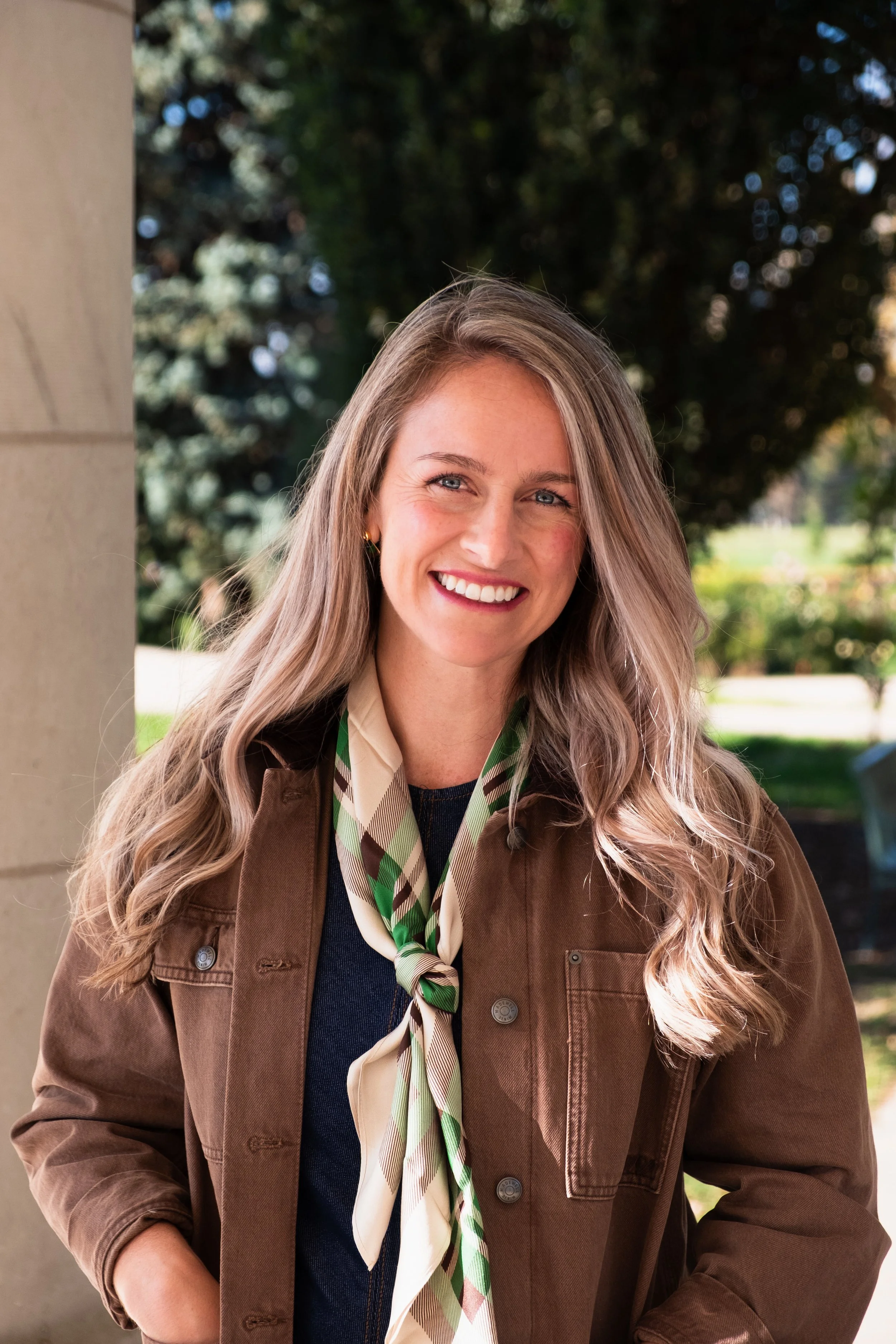 Smiling woman with long blonde hair wearing a brown jacket and patterned scarf outdoors