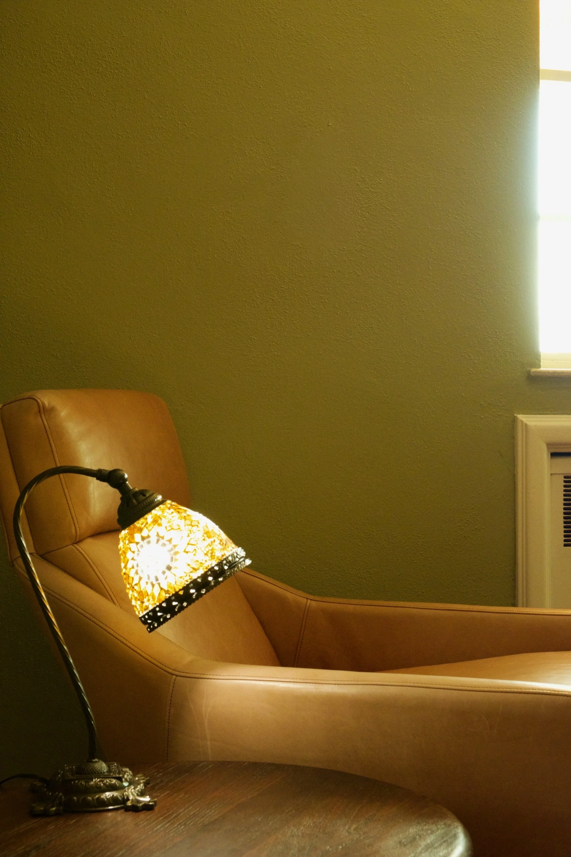 Cozy living room with a brown leather armchair, glowing mosaic table lamp, textured olive-green wall, window, and piano.