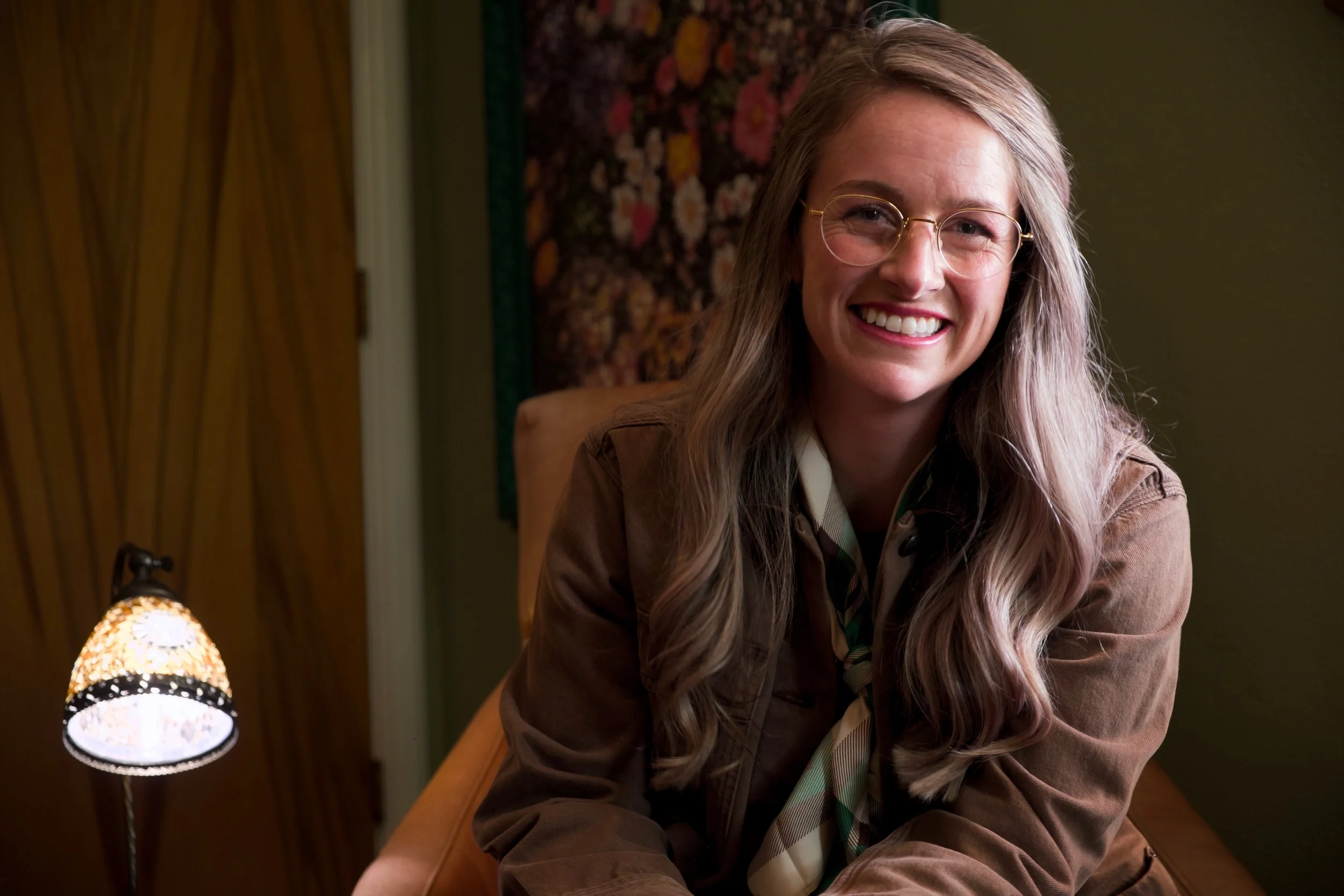 Smiling woman with long gray hair and round glasses sitting in a warmly lit room