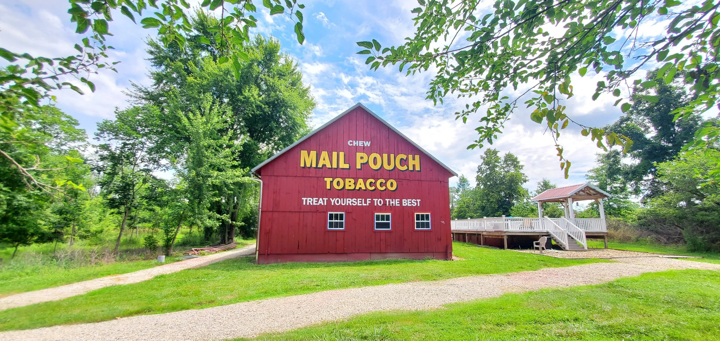 A red barn with white lettering advertising chew mail pouch tobacco, surrounded by green trees and grass on a sunny day.