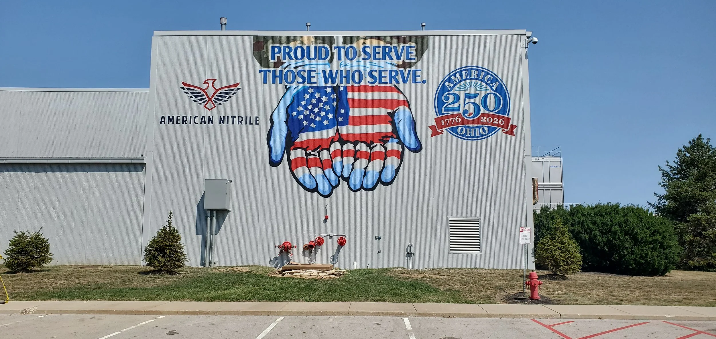 A large mural on the side of a building features a pair of hands painted with the American flag, with the text "PROUD TO SERVE THOSE WHO SERVE." included above. There are logos for American Nitrite and a 250th anniversary emblem for Ohio on the mural