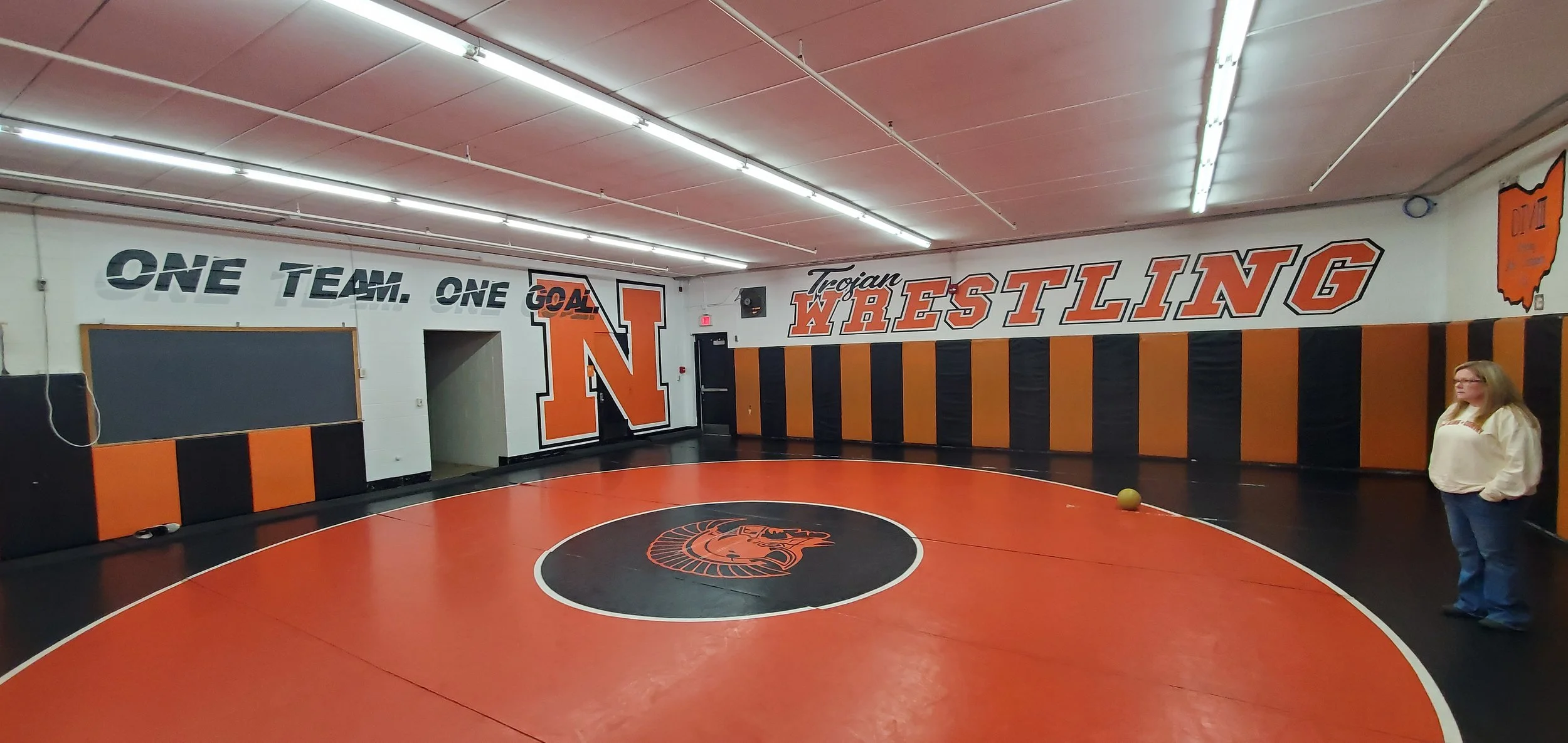 Wrestling gym with orange and black colors, wall murals displaying 'One Team. One Goal.' and 'Trojan Wrestling,' a painted 'N' logo, and a circle with a Trojan mascot in the center on the wrestling mat. There is a woman standing on the right side of 