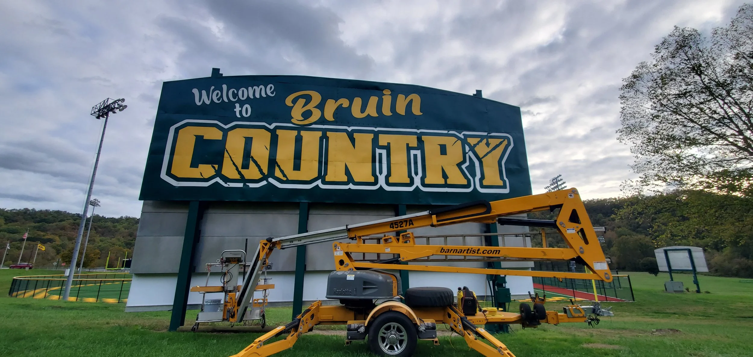 A large sign reads 'Welcome to Bruin Country' with a cloudy sky above. There are rolling hills, trees, football field lights, and some sports equipment in the background. In the foreground, a yellow lift is positioned beneath the sign.