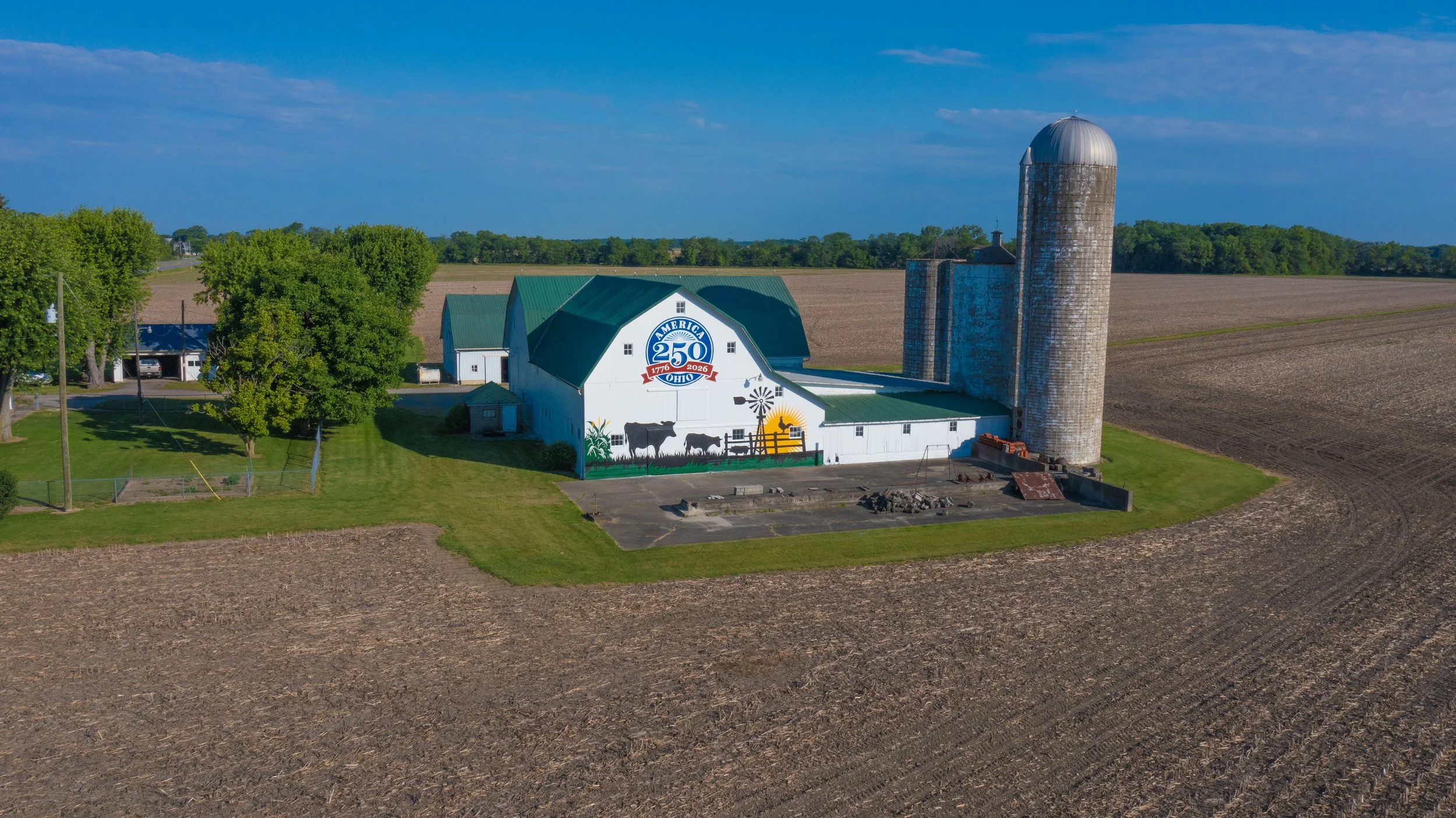 A farm building with a mural celebrating Ohio's 250th anniversary, featuring cows, a windmill, and crops, with silos nearby and farmland extending into the distance.