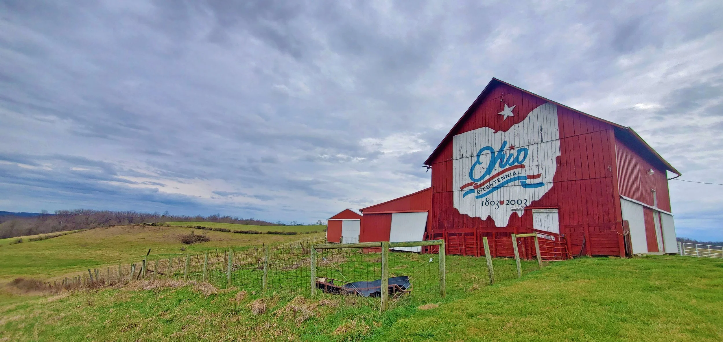 A red barn with a mural of the state of Ohio and the words 'Ohio Bicentennial 1803-2003' painted on it, standing in a rural green field with a cloudy sky overhead.
