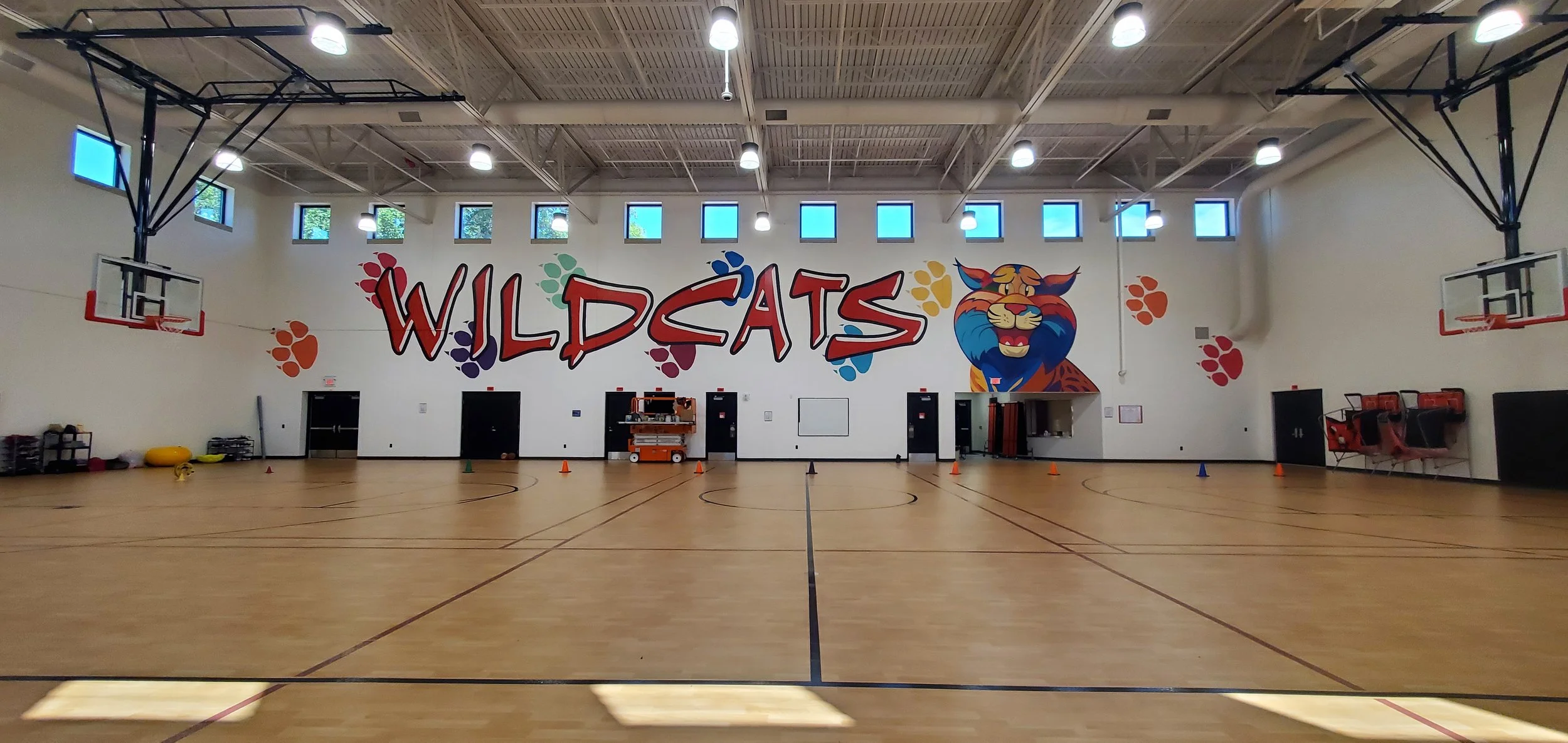 An empty indoor basketball court with a large colorful mural of a wildcat and the word 'WILDCATS' on the wall. The gym has high windows, ceiling lights, and sports equipment along the walls.